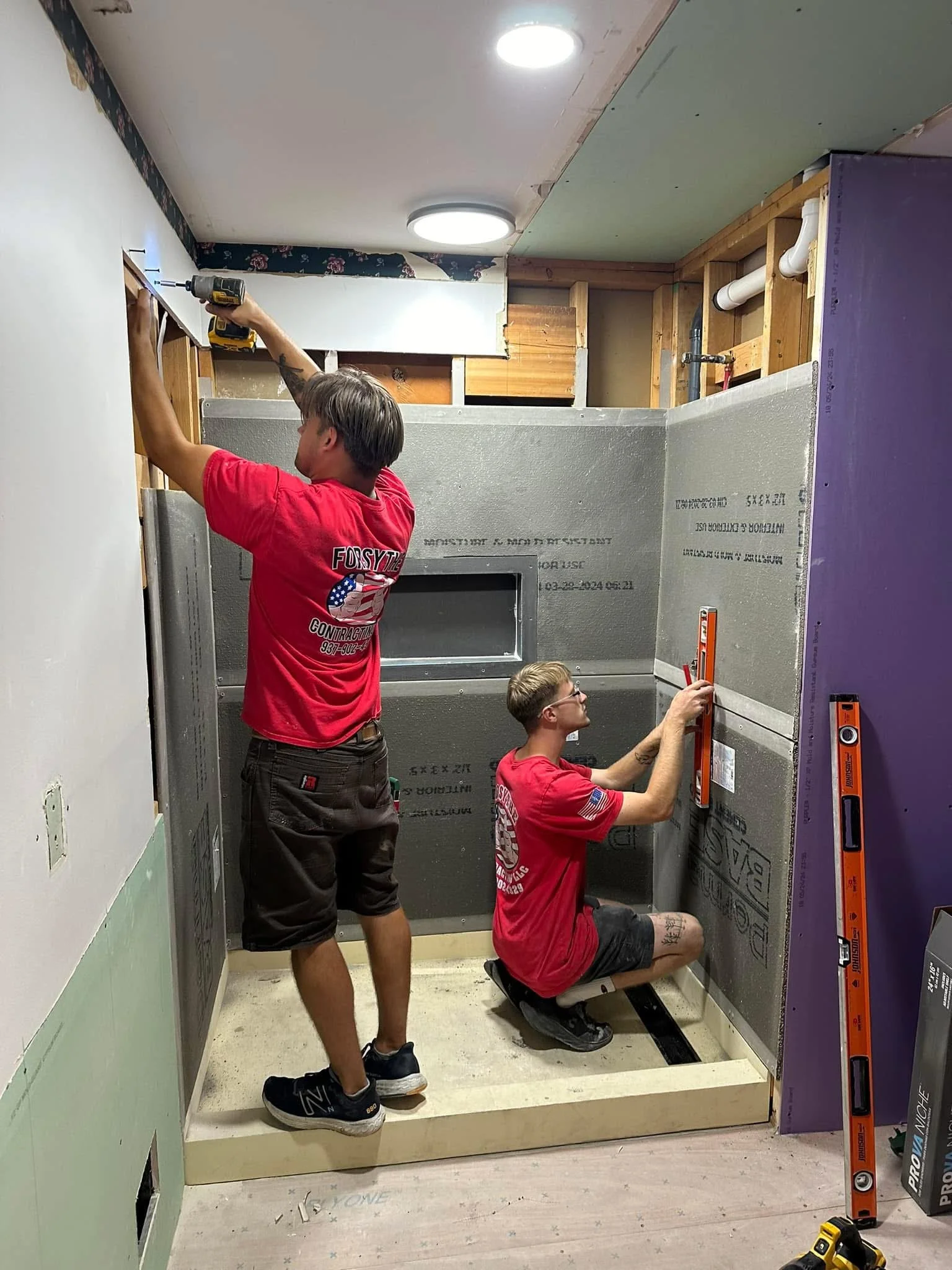 Two men working on a construction project inside a house, with one using a power drill and the other measuring with a level.