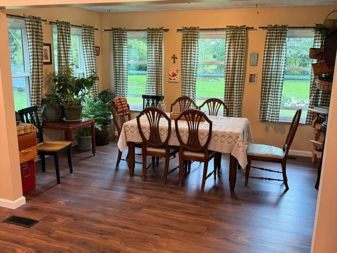 Dining room with wooden dining table, six chairs, and checkered curtains on windows showcasing green outdoor scenery.