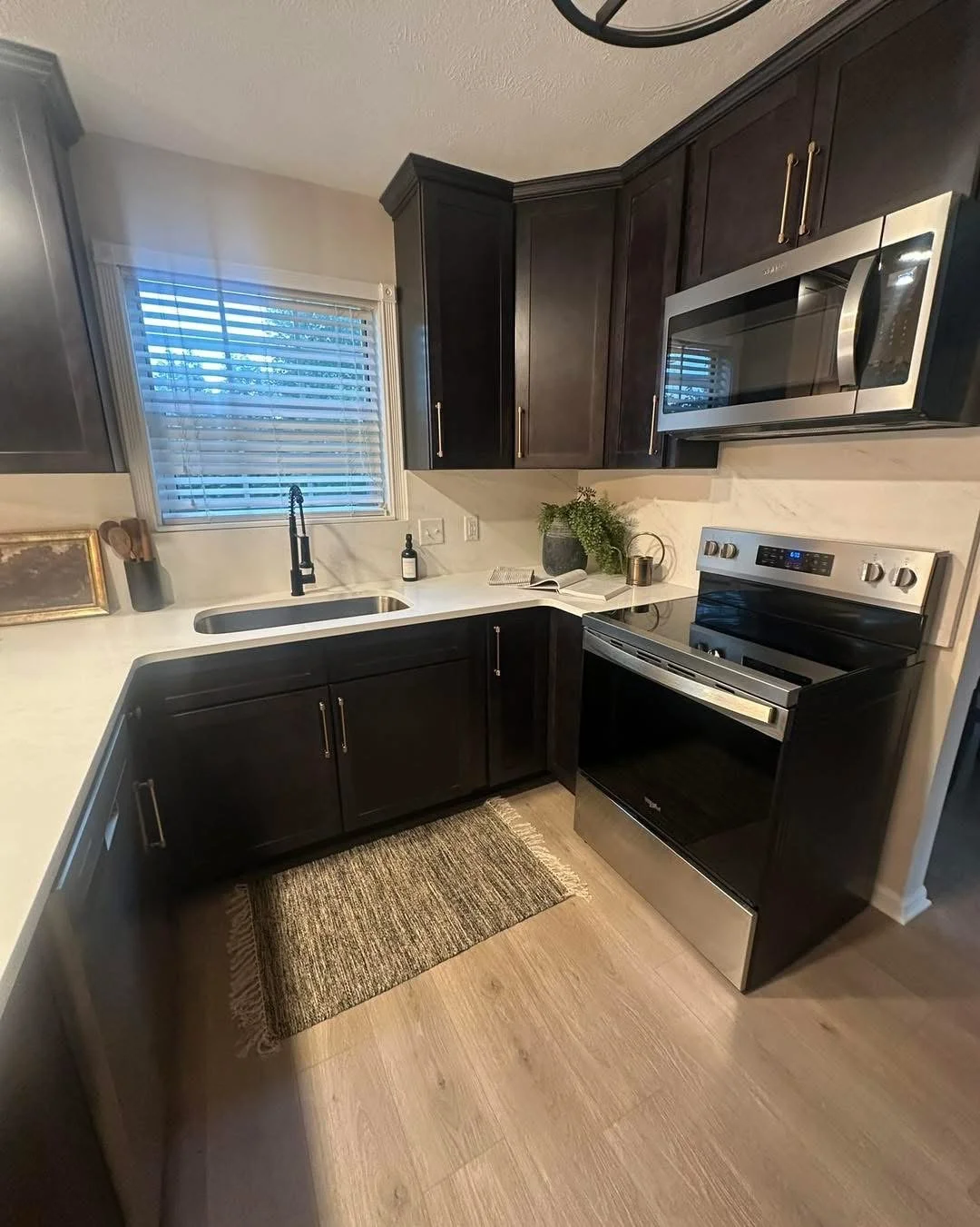 Kitchen with black cabinets, white countertops, a window with blinds, stainless steel microwave and oven, black faucet, and some plants on the counter.