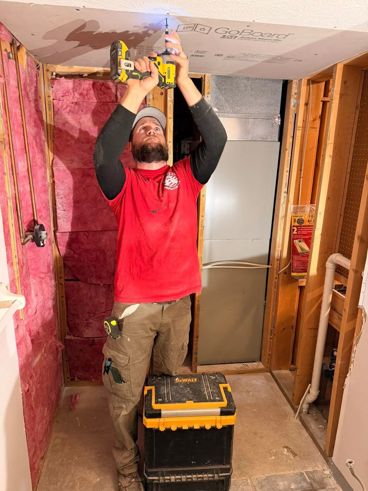 A man installing drywall in a partially framed room with pink insulation and exposed wooden studs, using a cordless drill, with a toolbox on the floor