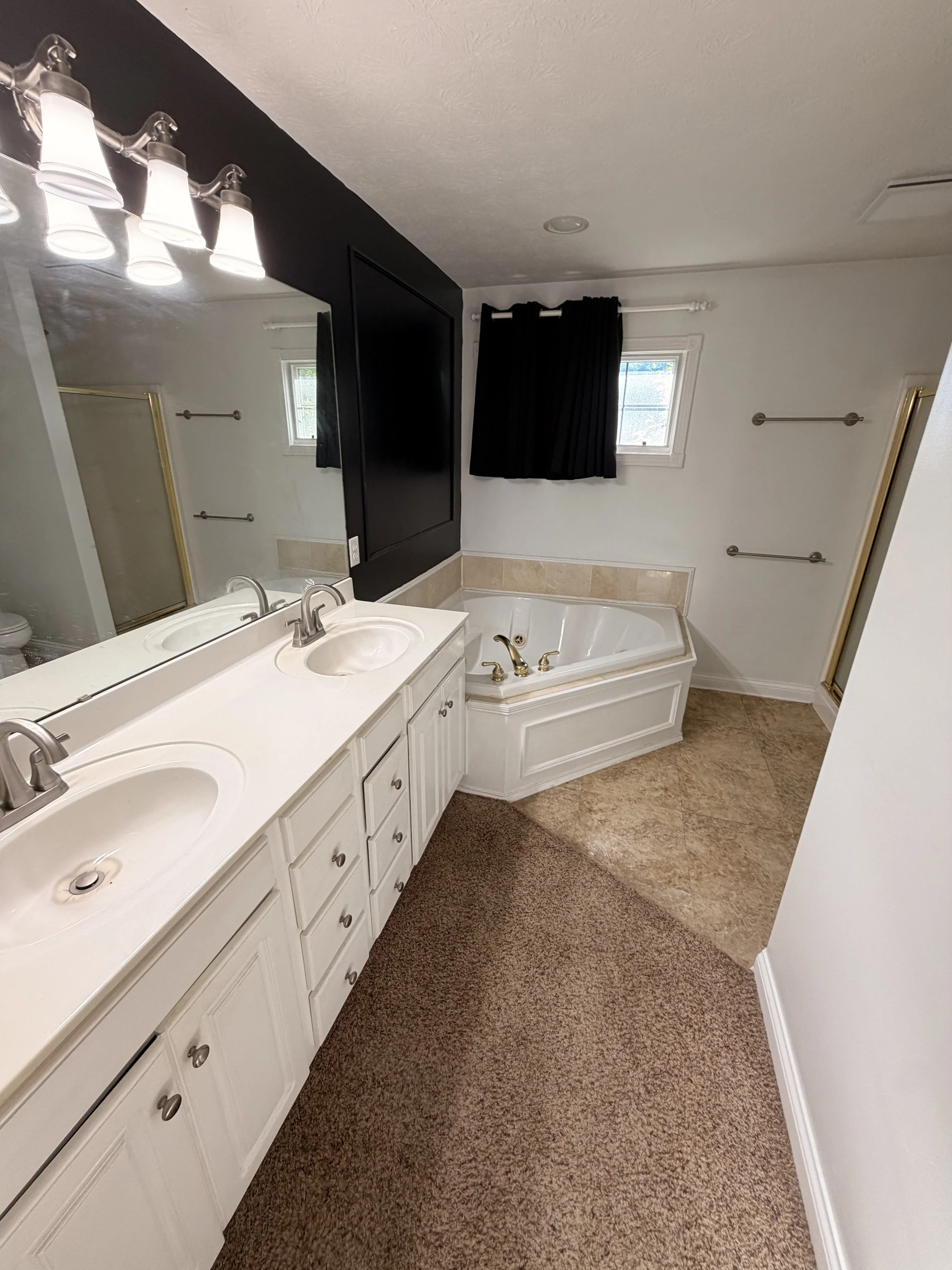 Bathroom with white double sink vanity, large mirror, beige tile bathtub surround, black curtain, and brown carpeted floor.