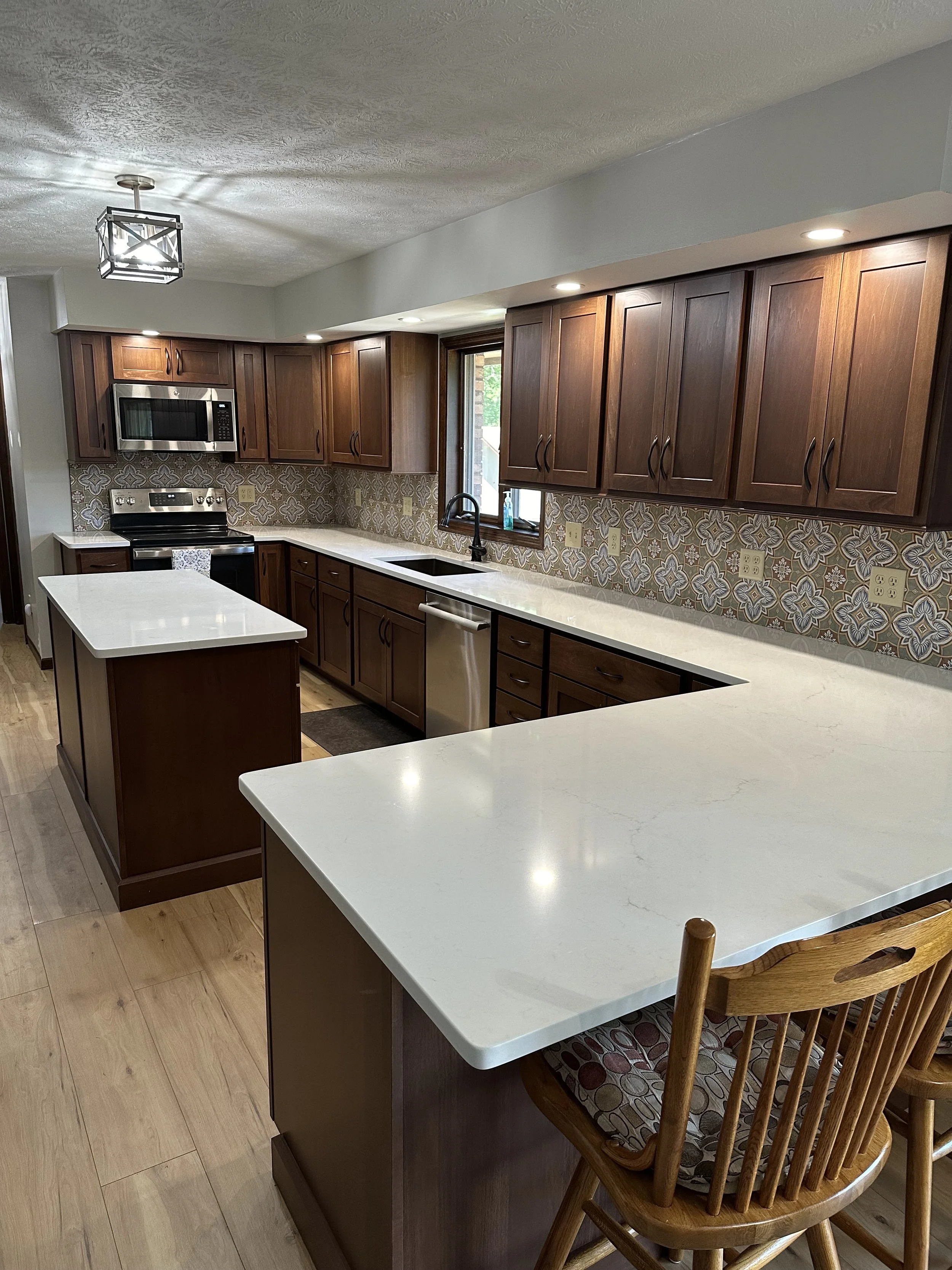 Kitchen with wooden cabinets, a white countertop island, and patterned backsplash tile, with a window above the sink and a dining chair in the foreground.
