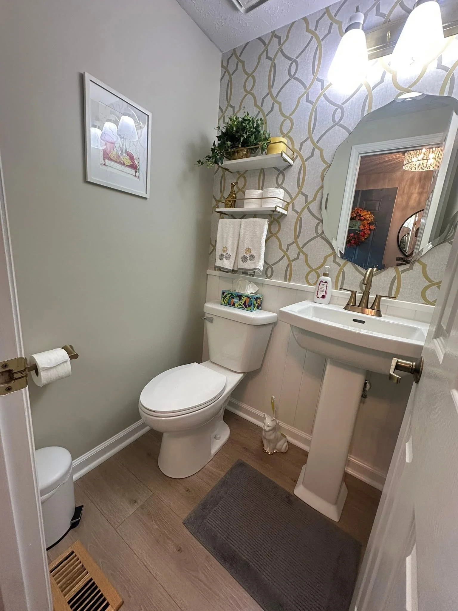 A small bathroom with a toilet, a sink with a gold faucet, a mirror, and wall decor. There is a shelf with a plant and towels, a framed picture on the wall, and a gray rug on wood flooring.