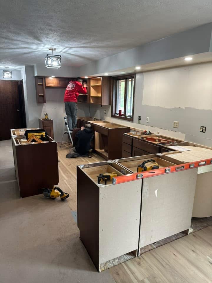 Two people installing kitchen cabinets in a home under renovation, with tools and unfinished cabinetry visible.