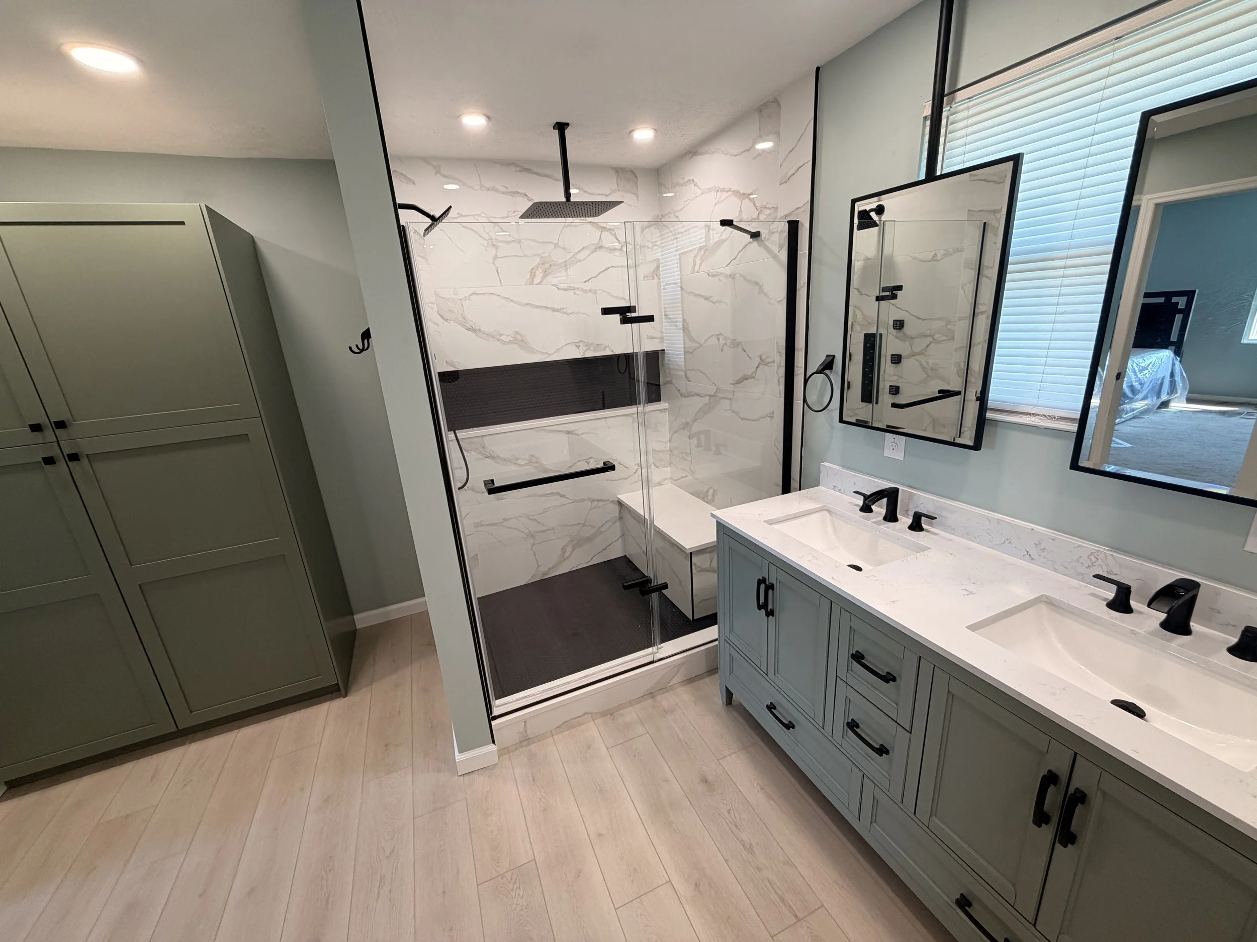 Modern bathroom with a walk-in shower featuring a black rain showerhead and marble wall tiles, a bathroom vanity with a white countertop, black fixtures, and mirrors, window with blinds, and light wood flooring.