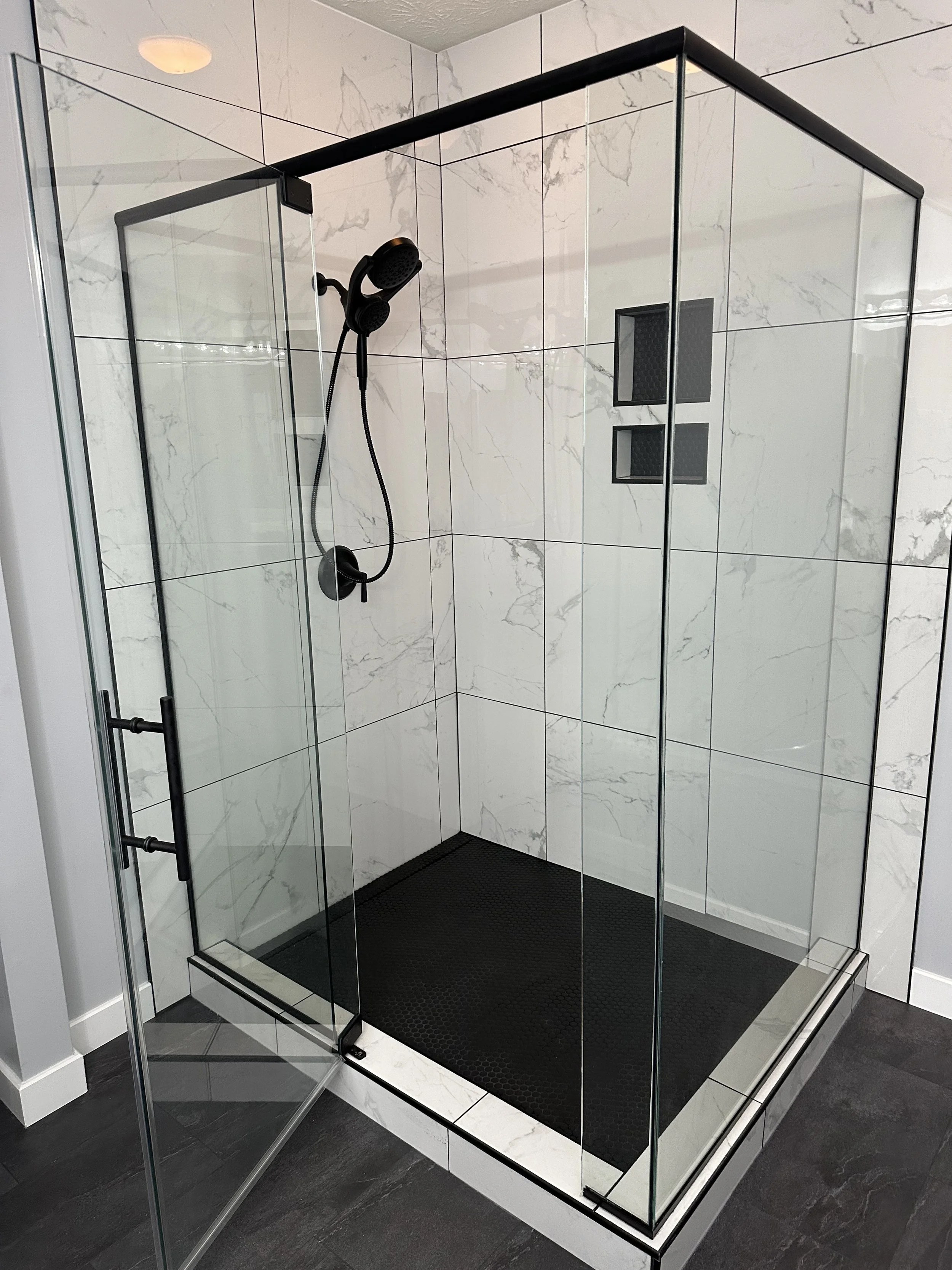 A modern shower stall with glass walls, a black framed enclosure, black shower fixtures, and black square tile shelves built into the white marble tiled wall.