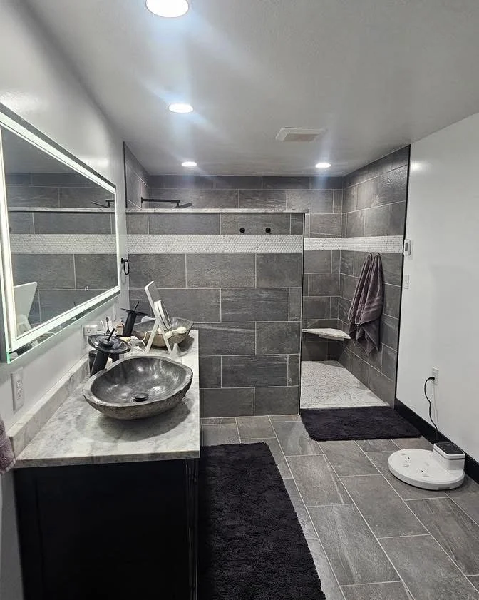 Modern bathroom with gray tiled walls and floor, black bathroom cabinet with gray marble countertop, stone vessel sink, large mirror, walk-in shower with bench and towel hanging, black rug, and a robotic vacuum on the floor.