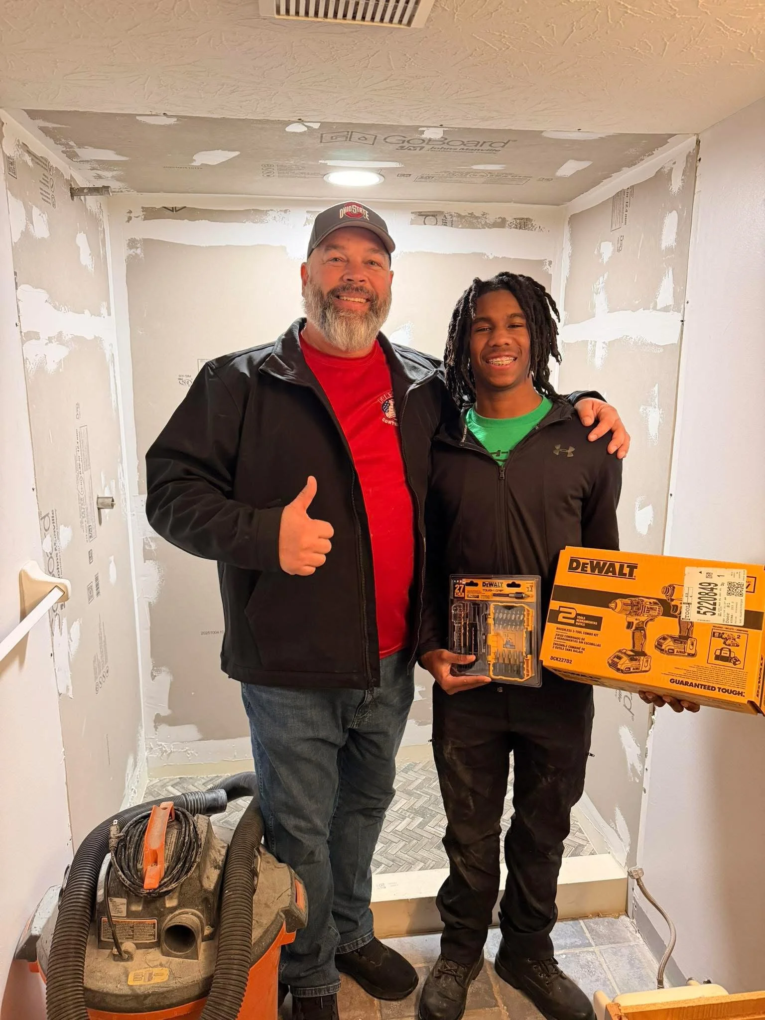 Two men standing in a partially renovated room, smiling, with construction tools and materials around them, one holding a box of DeWalt power tools.