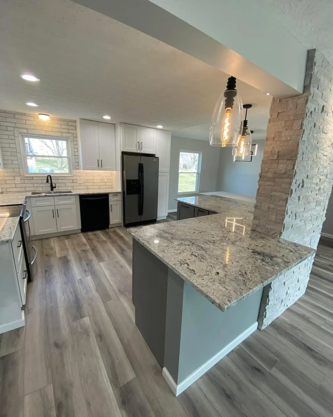 Modern kitchen with white cabinets, black appliances, granite countertops, wood-style flooring, stone accent wall, and pendant lighting.