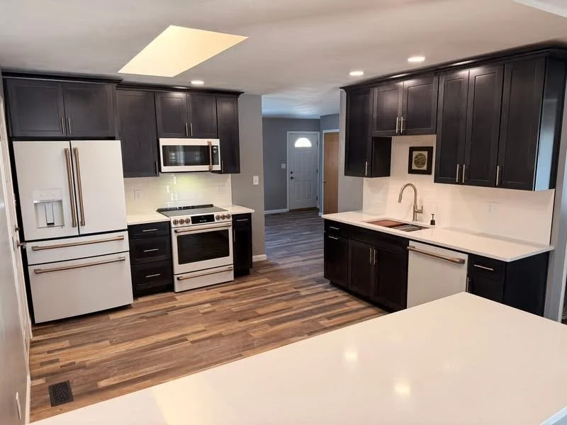 Modern kitchen with dark cabinets, white countertops, and white appliances including a refrigerator, stove, microwave, and dishwasher. Wooden flooring and a partially visible dining area or adjacent room in the background.