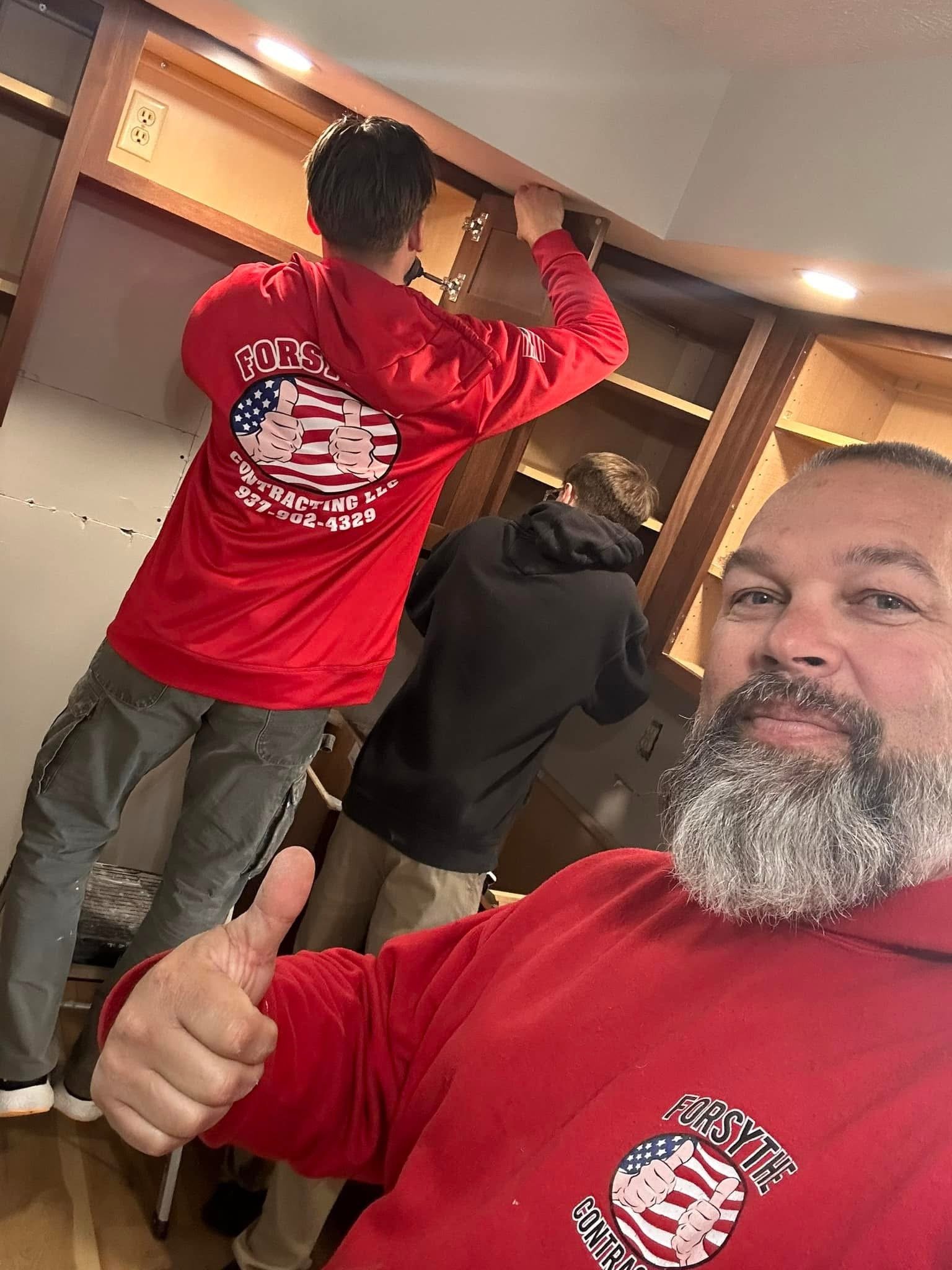 A man giving a thumbs-up, wearing a red sweatshirt with a logo that says 'FORSY THE CONTRA' and an American flag design. In the background, people are working on a wooden cabinet or shelving unit, with one person using a screwdriver.