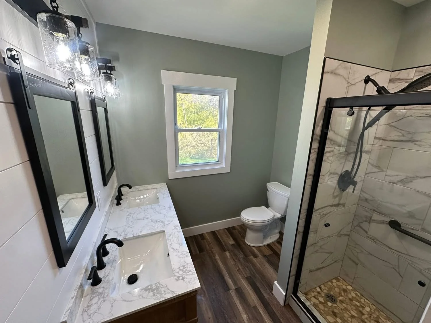 Modern bathroom with two sinks with black faucets, marble countertops, and framed mirrors, a window above the toilet, a glass-enclosed shower with marble tiles and a pebble floor, and wood flooring.