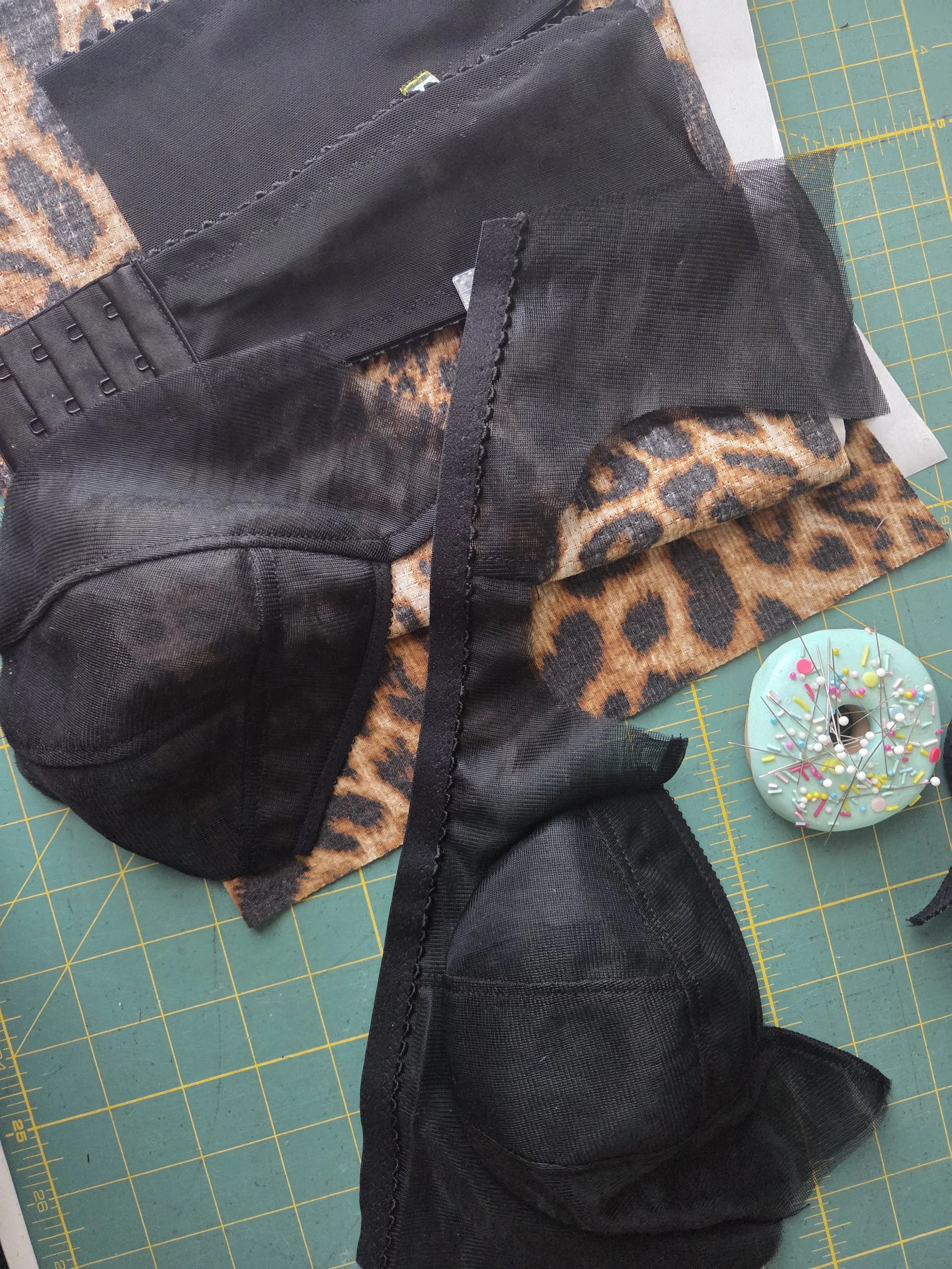 Black mesh fabric and a strip of black elastic lying on a cutting mat with a leopard print fabric underneath, a magnetic donut pincushion with pink and white sprinkles off to the side.