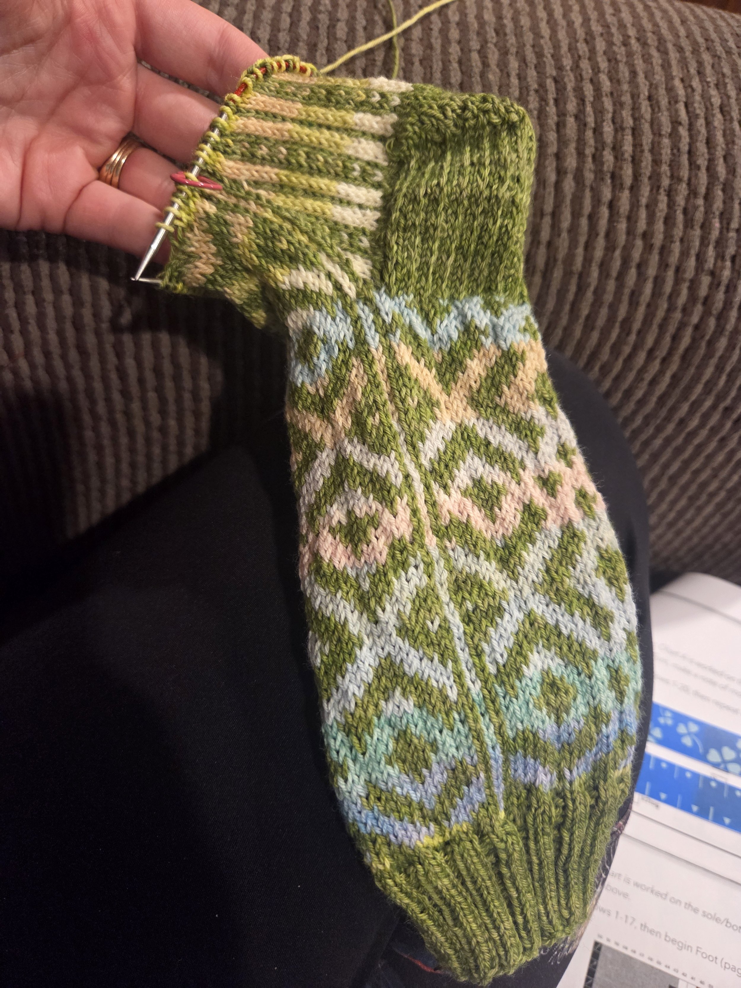 Close-up of a colorful knitted sock with various shades of green, beige, light blue, and some other colors, being worked on with knitting needles.