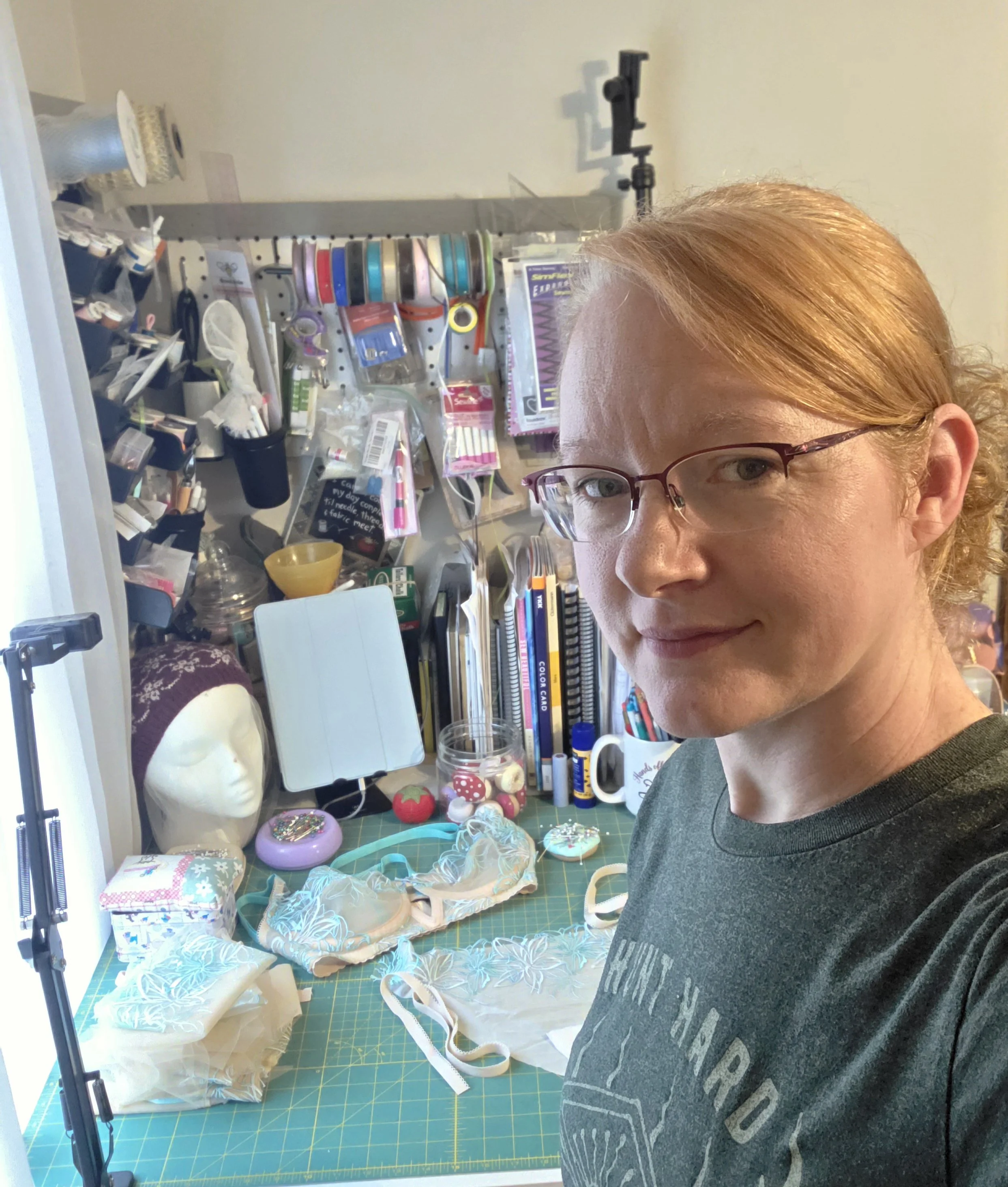 A person with red hair and glasses standing in a craft room with a green cutting mat and various sewing and craft supplies, including fabric, ribbons, and decorative items, on a table behind them.