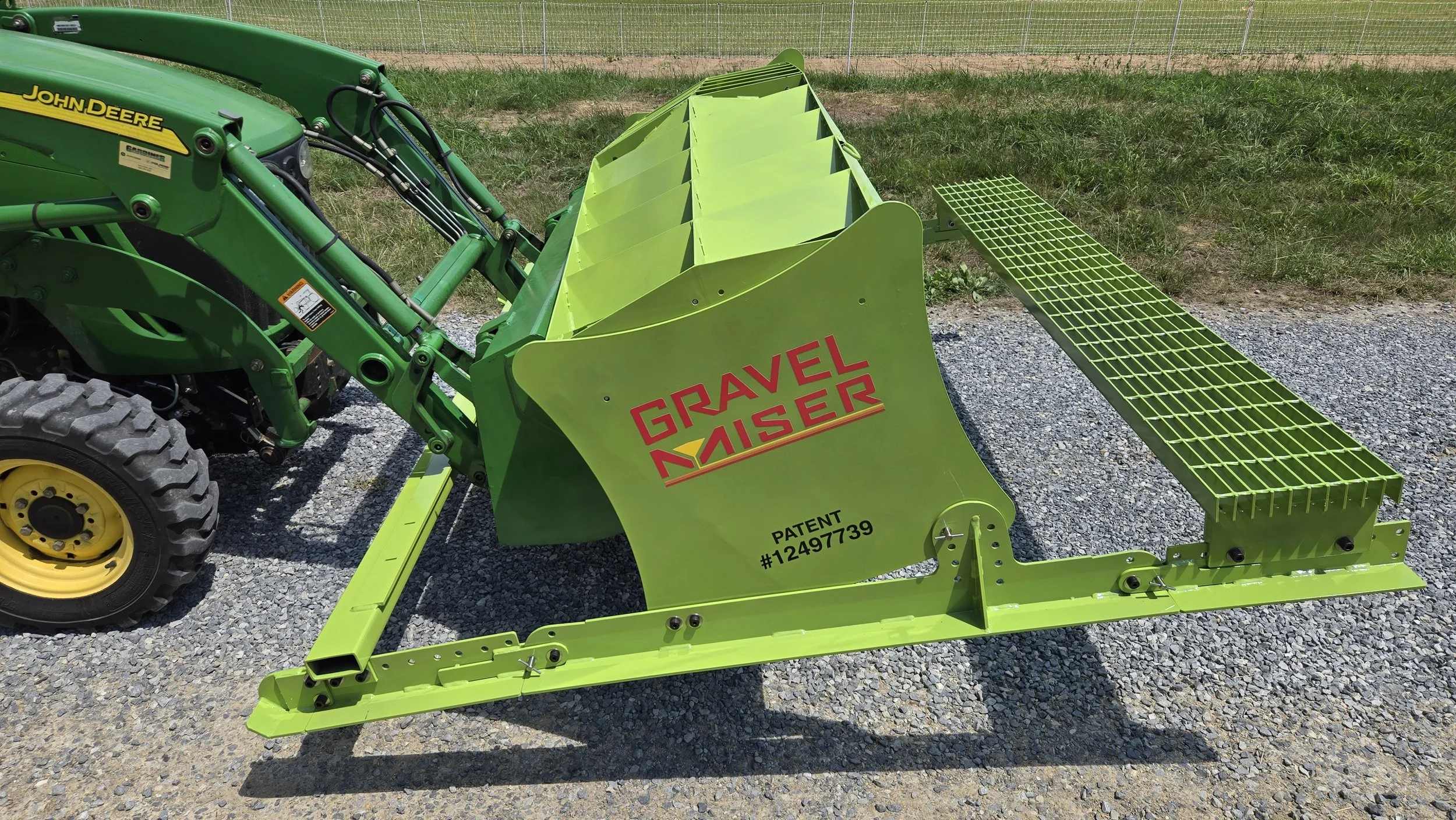 Green John Deere tractor with a yellow wheel, attached to a lime green gravel travel separator with red and black text, parked on a gravel surface with a grassy field and fence in the background.