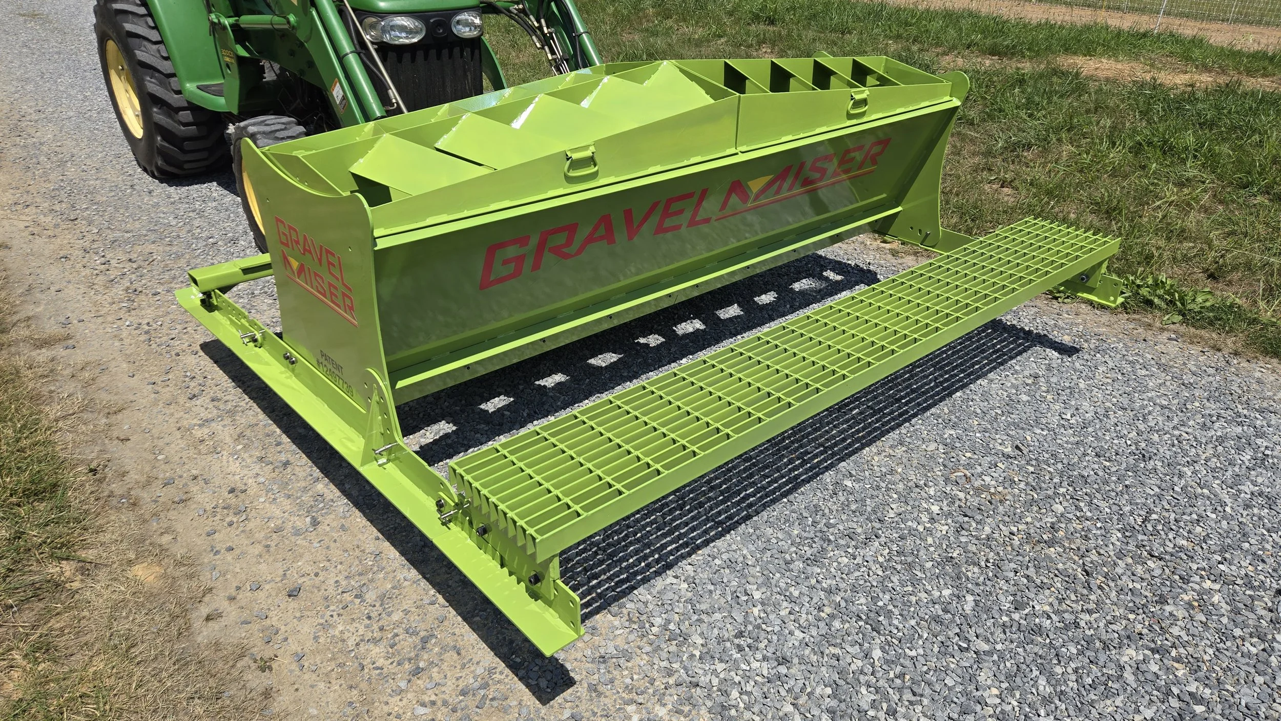 Green gravel cleaner attachment labeled 'Gravel Master' attached to a tractor on a gravel path next to a grassy field.