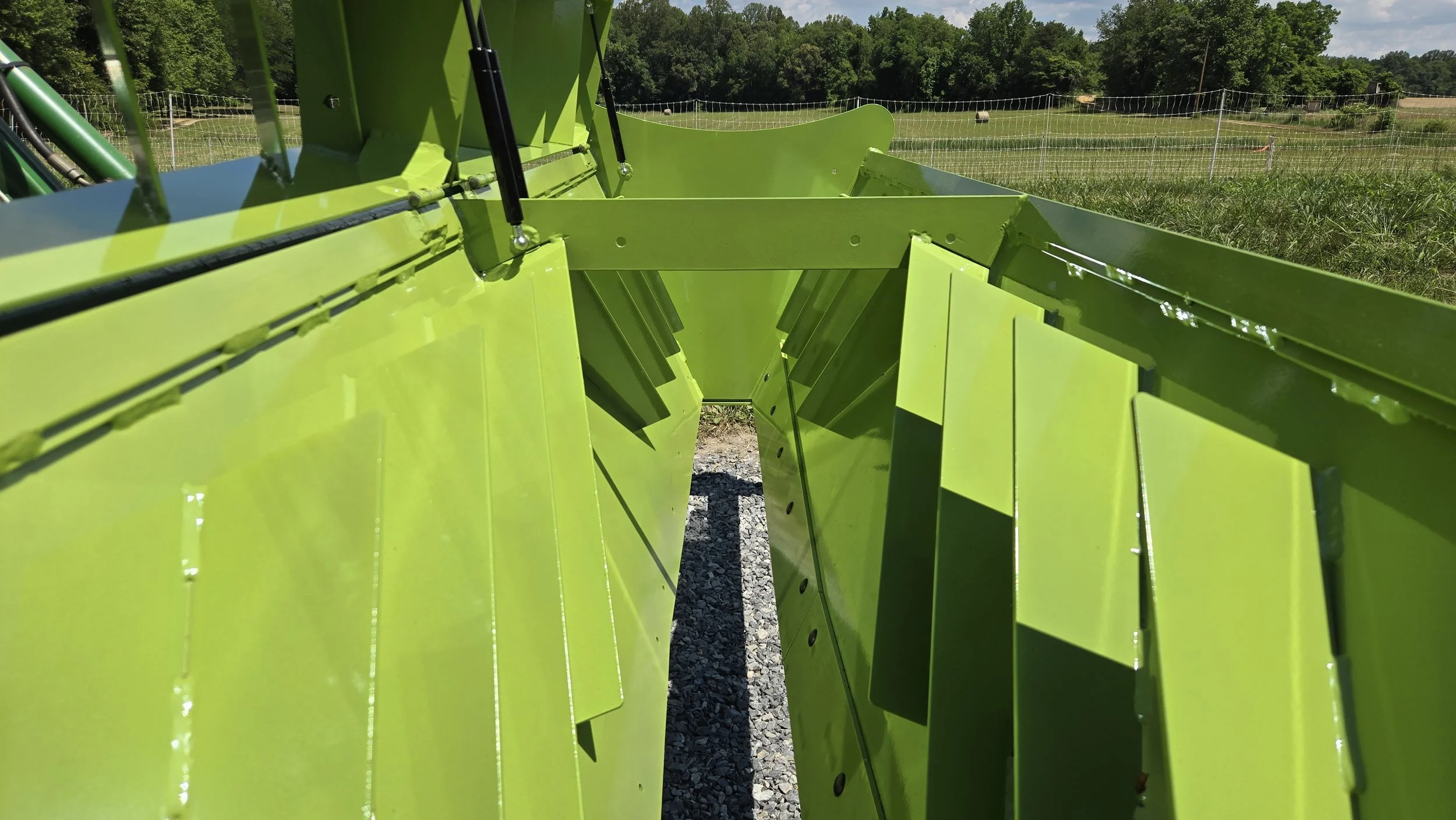 Close-up of a large green agricultural machine or equipment outdoors on a grassy field with trees and a blue sky in the background.