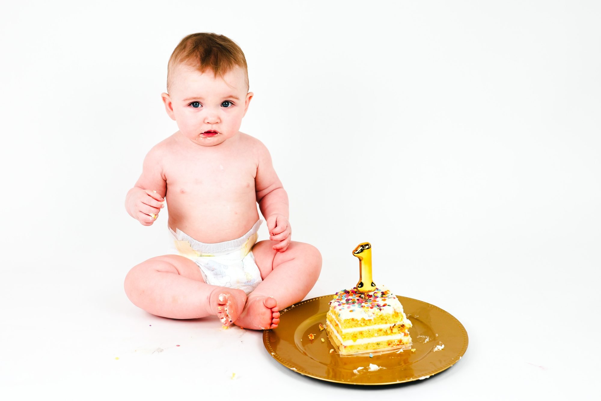 One-year-old baby sitting in a diaper beside a small cake with a gold number one candle on a gold plate, cake smash portrait