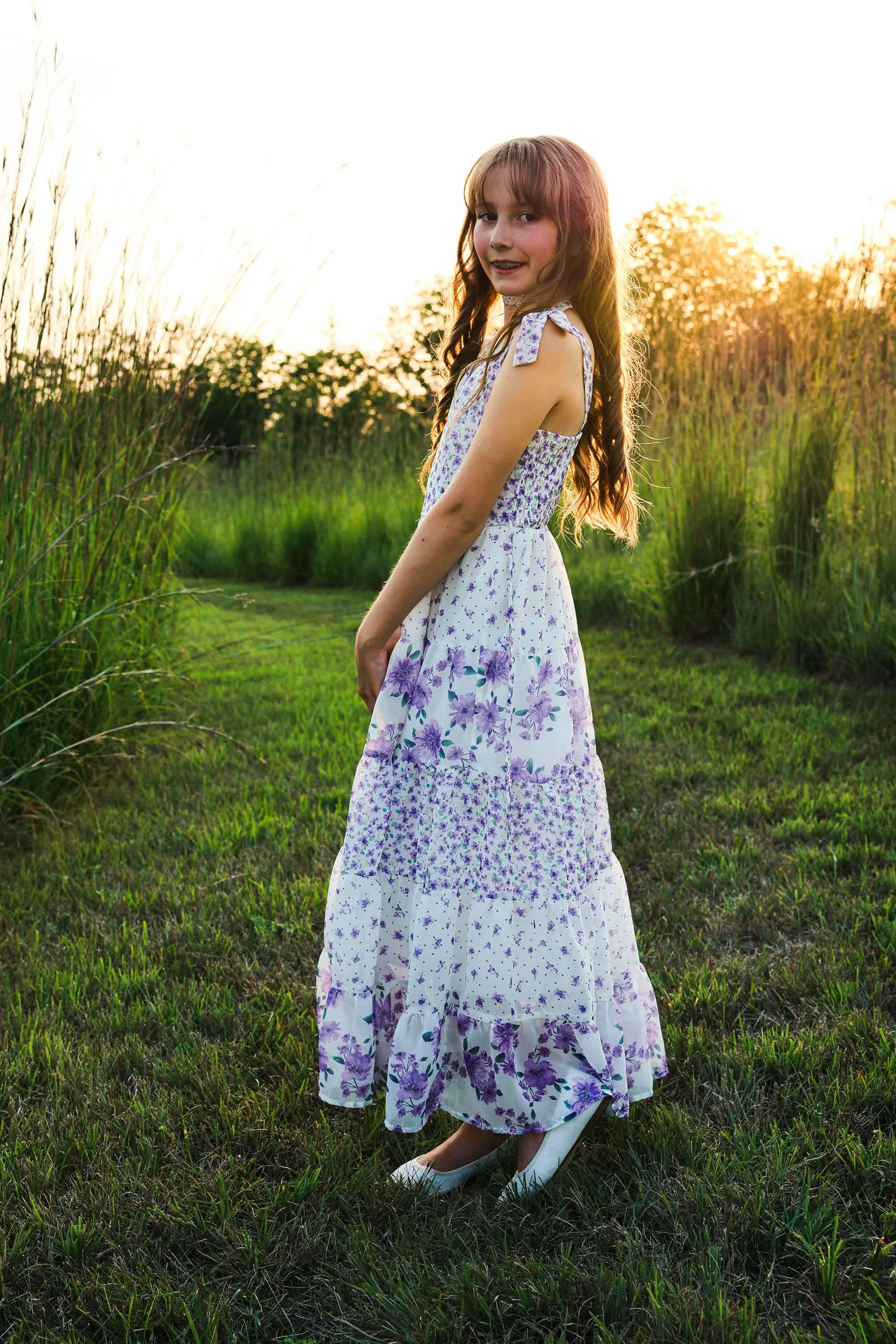 The girl glancing back over her shoulder in a white and purple floral tiered maxi dress, backlit by the sunset.