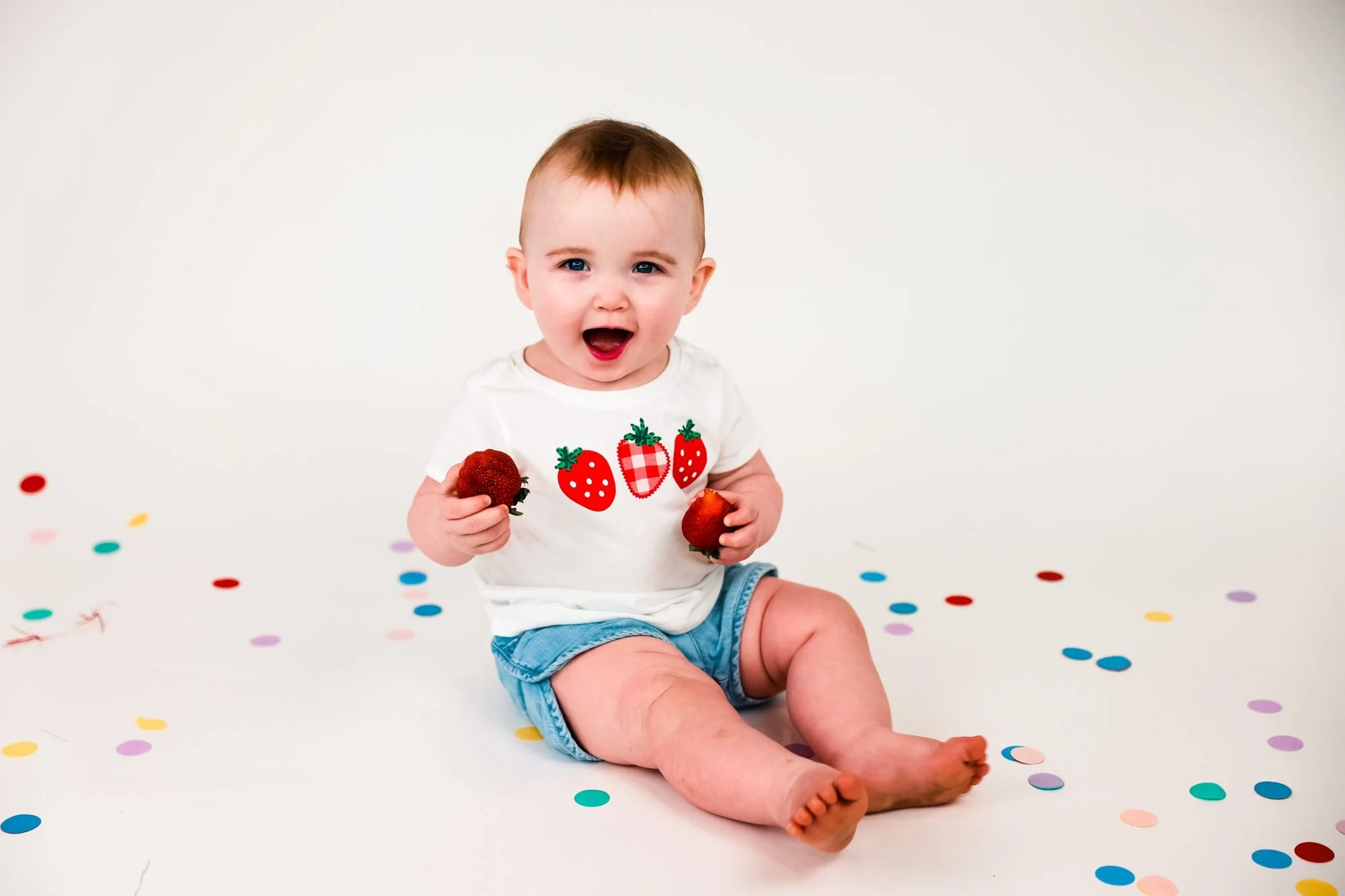 Smiling baby in a strawberry shirt and denim shorts sitting on a white floor scattered with colorful confetti, holding strawberries