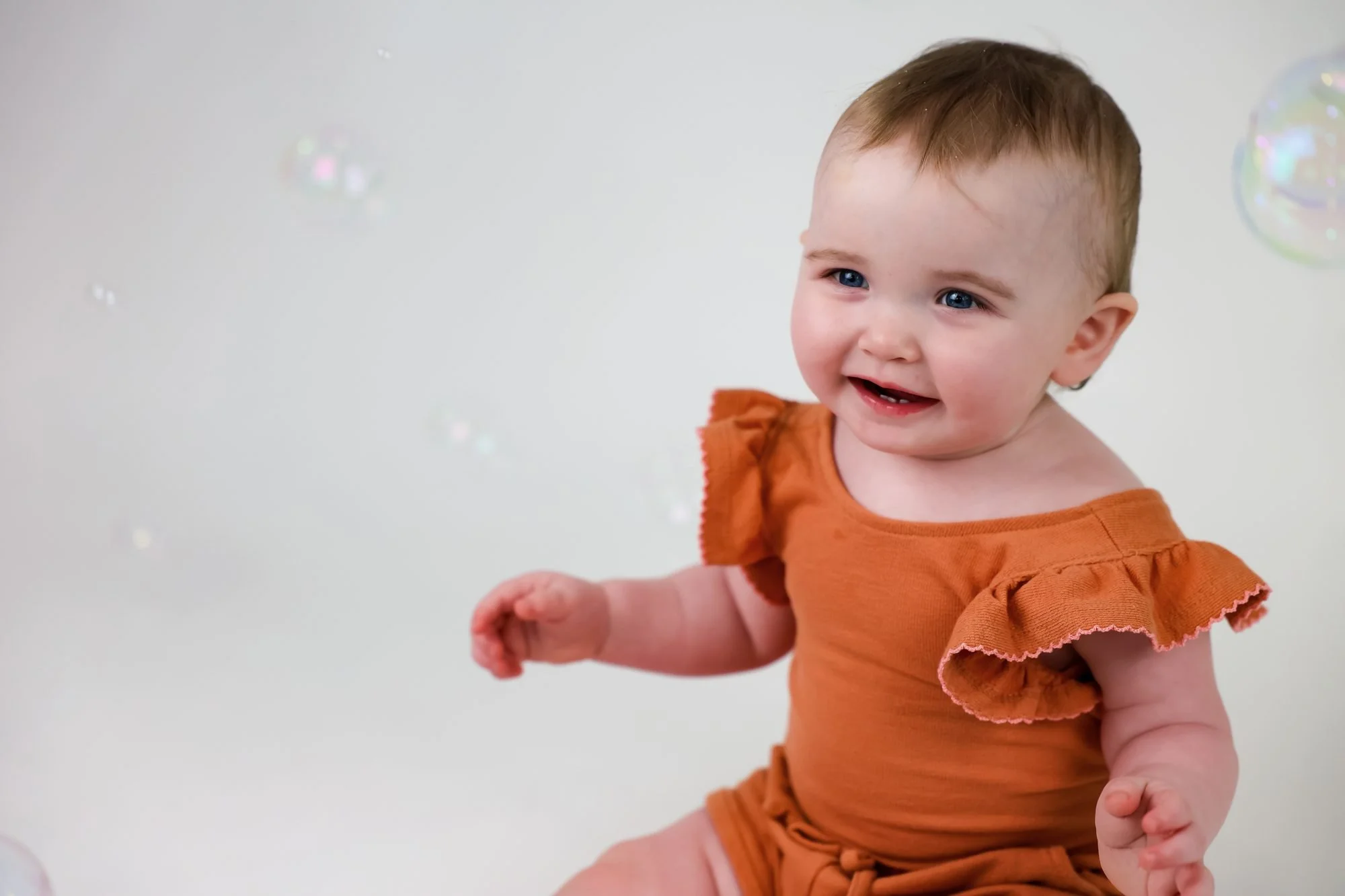 Smiling baby in a rust-orange ruffled top with soap bubbles floating in the soft background