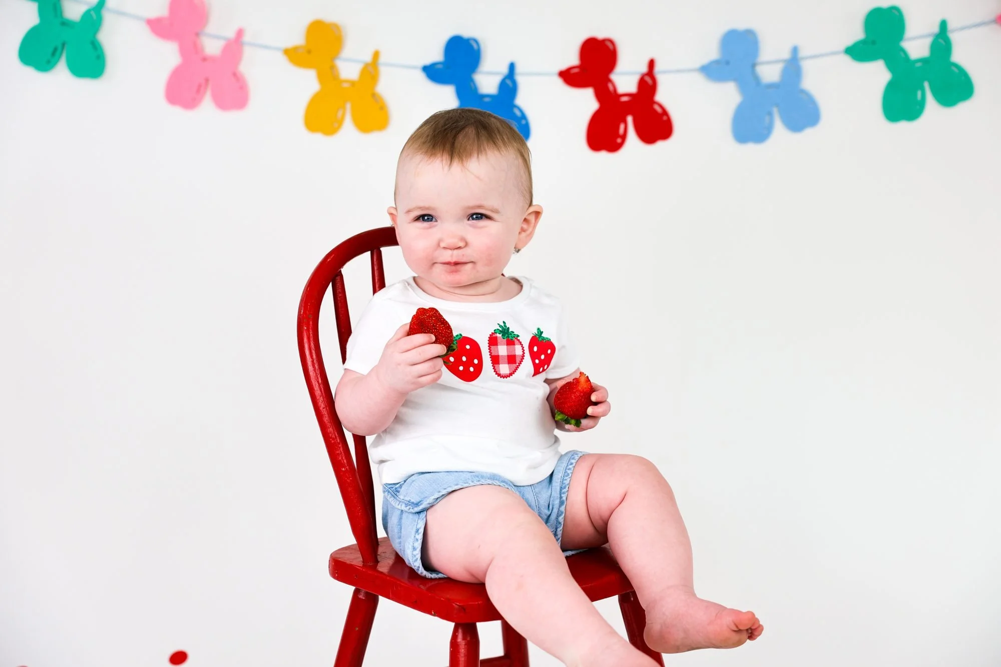 Baby in a white strawberry shirt and denim shorts sitting on a red wooden chair, holding a strawberry, in front of a colorful balloon-dog garland