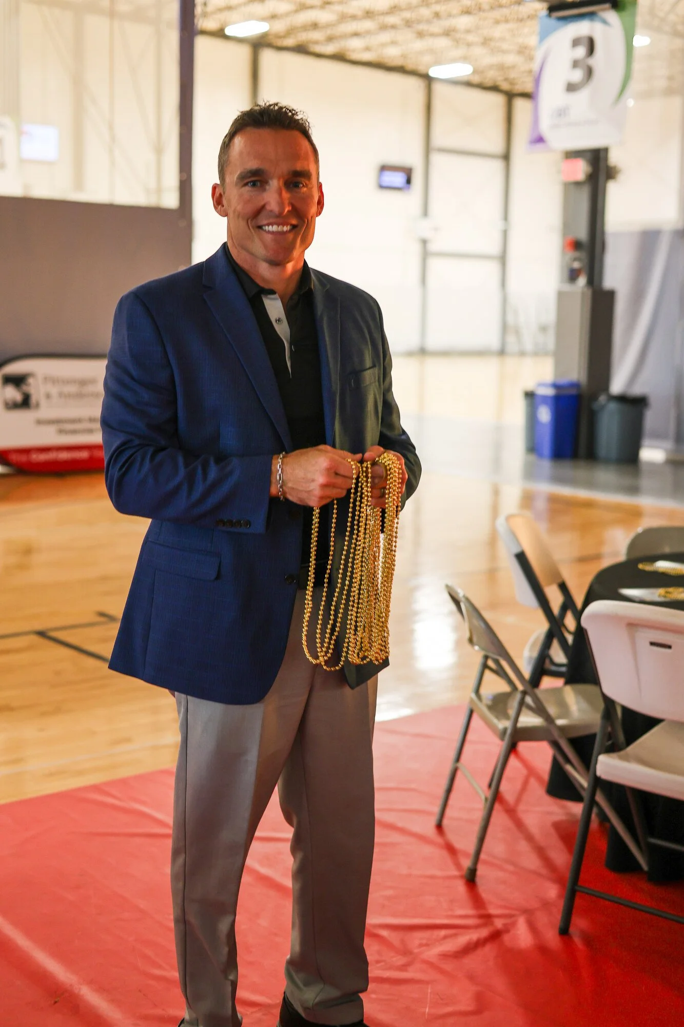 A man in a blue blazer holding strands of gold beads in a gym with a red floor.