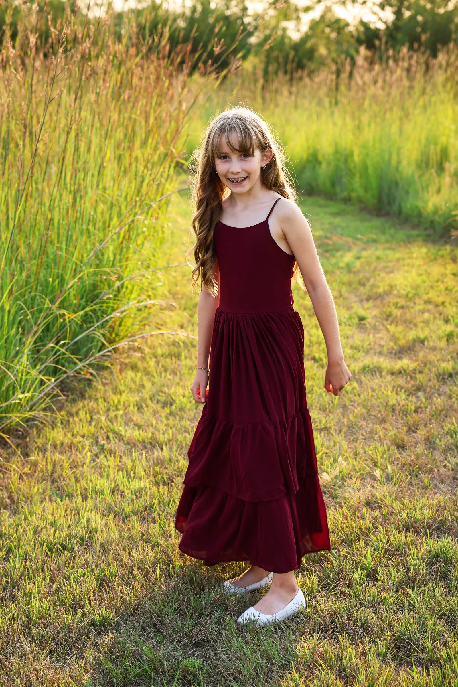A smiling girl in a burgundy tiered maxi dress standing in tall grass at golden hour.