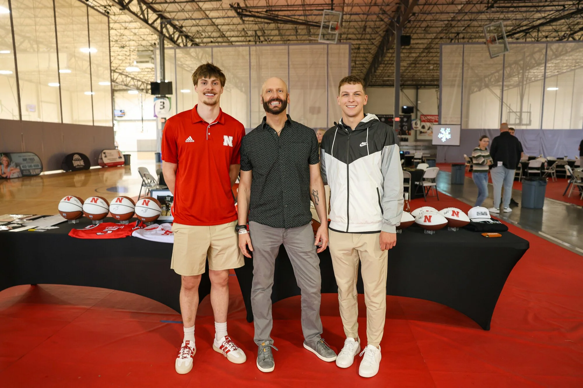 Three men standing in front of a table of signed Nebraska items, one wearing a red Nebraska shirt.