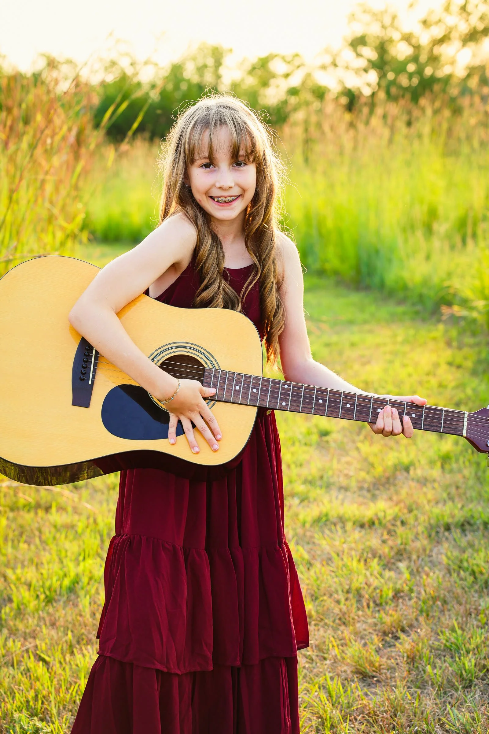 The girl in the burgundy dress holding an acoustic guitar in a grassy field at sunset.