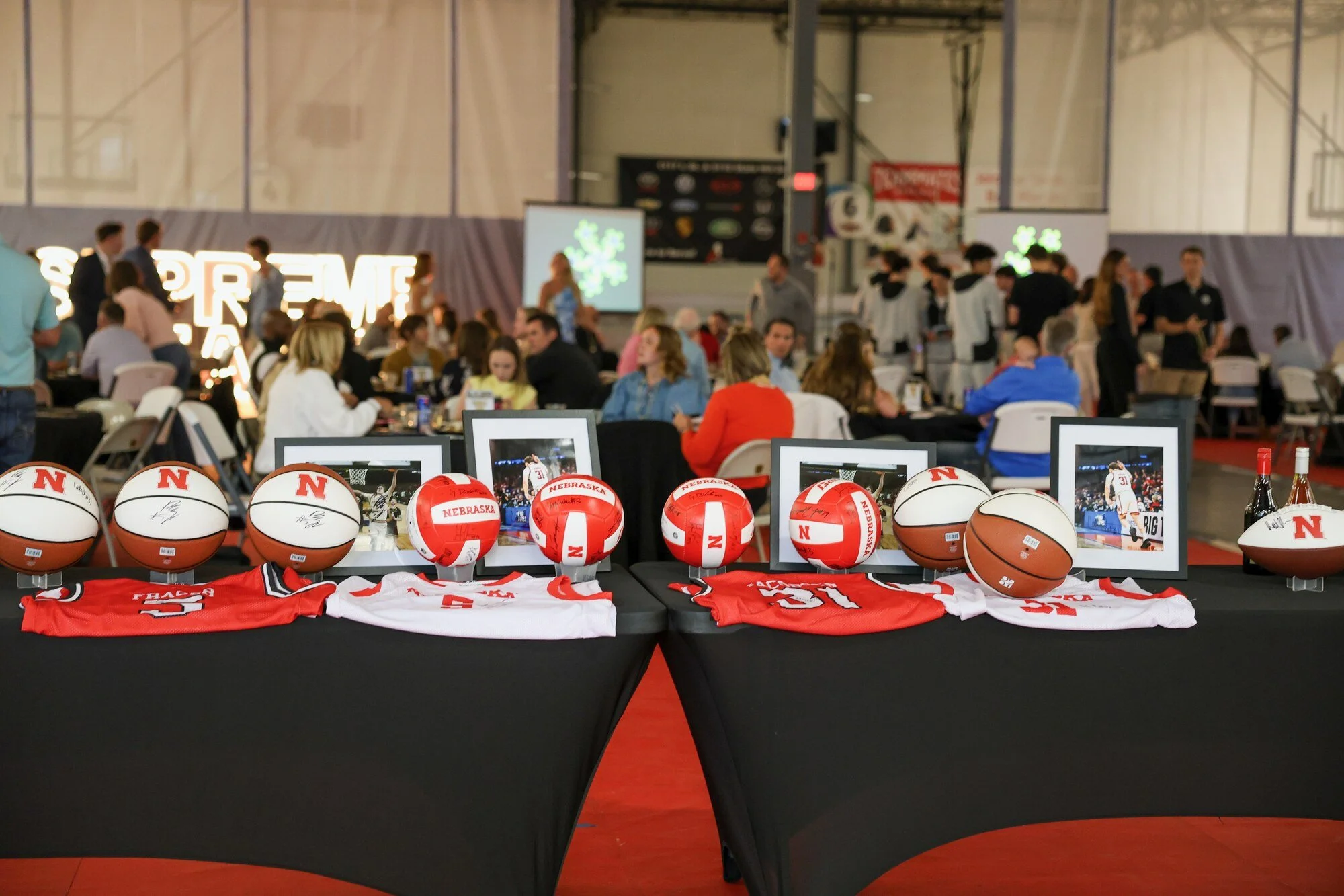 Auction display tables with signed Nebraska basketballs, volleyballs and jerseys and framed photos, with guests seated at tables behind.