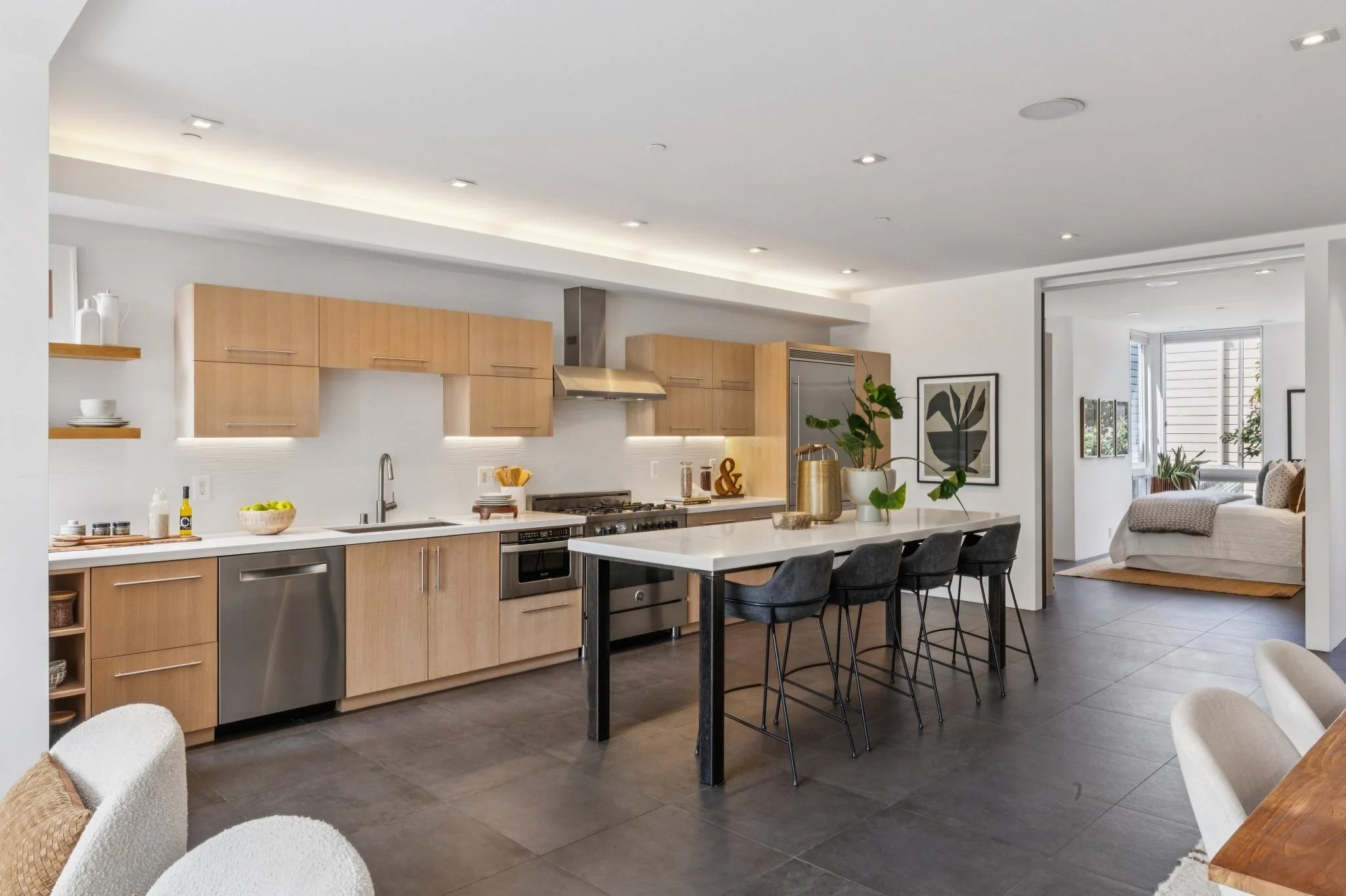 Open-concept modern kitchen with light wood cabinets, stainless steel appliances, and a white countertop island with black chairs, adjacent to a living room with large windows and framed artwork.