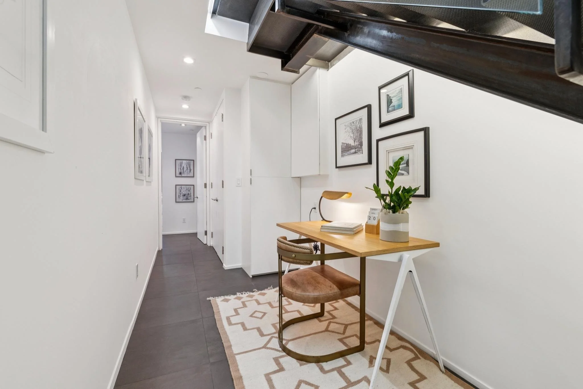 Interior of a modern home with white walls, framed artwork, a small wooden desk with a lamp, books, and a plant. A chair with a brown cushion is in front of the desk, and a patterned rug is on the dark flooring. The hallway leads to a closed door, wi