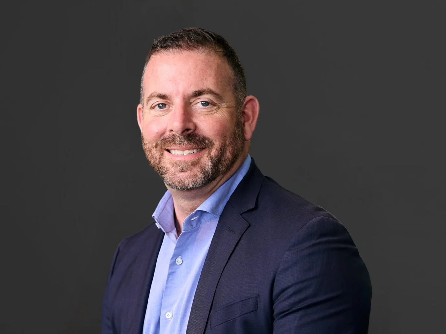 A professional headshot of a smiling man with short brown hair and a beard, wearing a dark suit jacket and light blue shirt, against a dark gray background.