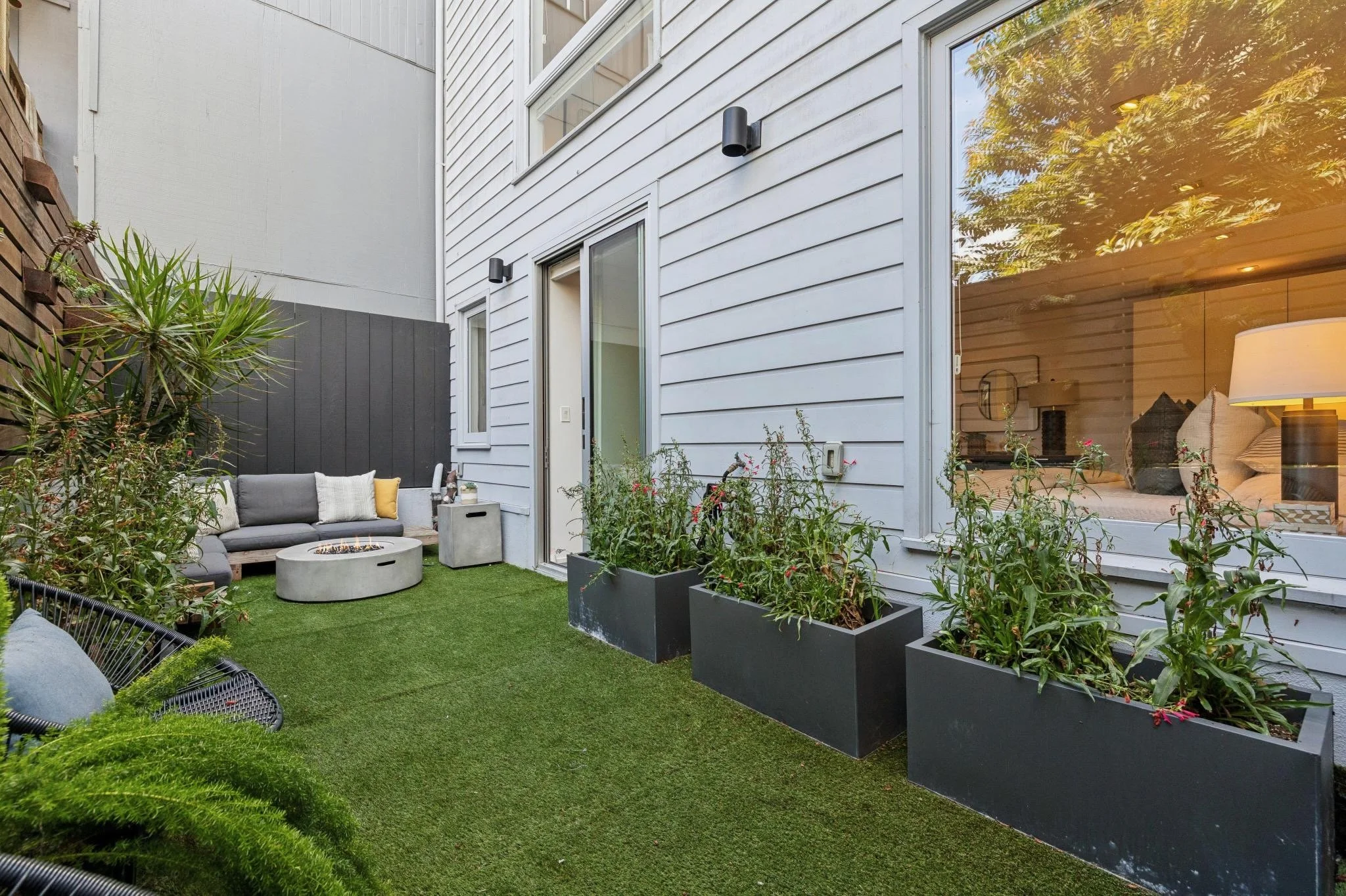 A backyard patio with outdoor seating, a fire pit, potted plants, and a view through a large window into a living room with a sofa and lamp.