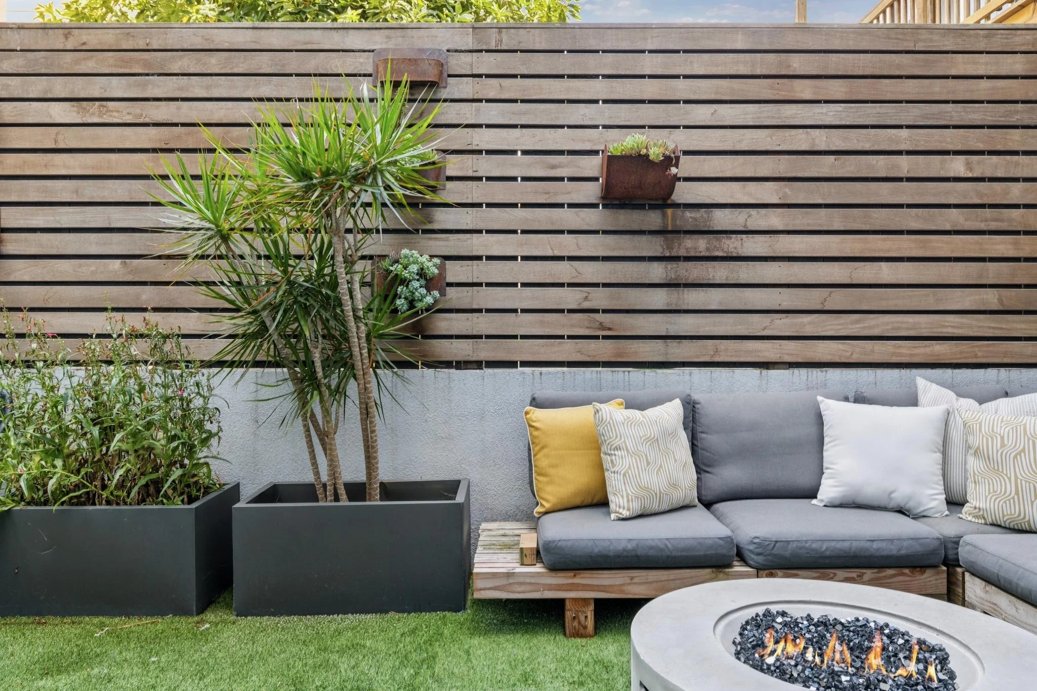 Outdoor patio area with a wooden slat fence, a gray sectional sofa with yellow and patterned pillows, potted plants including a tall tree and succulents, and a fire pit with glowing embers.