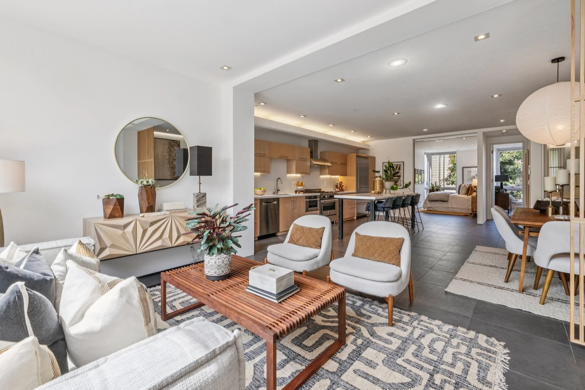 Open-concept living room and kitchen with modern decor, white walls, wooden accents, large windows with outdoor views, and various seating and decorative elements.