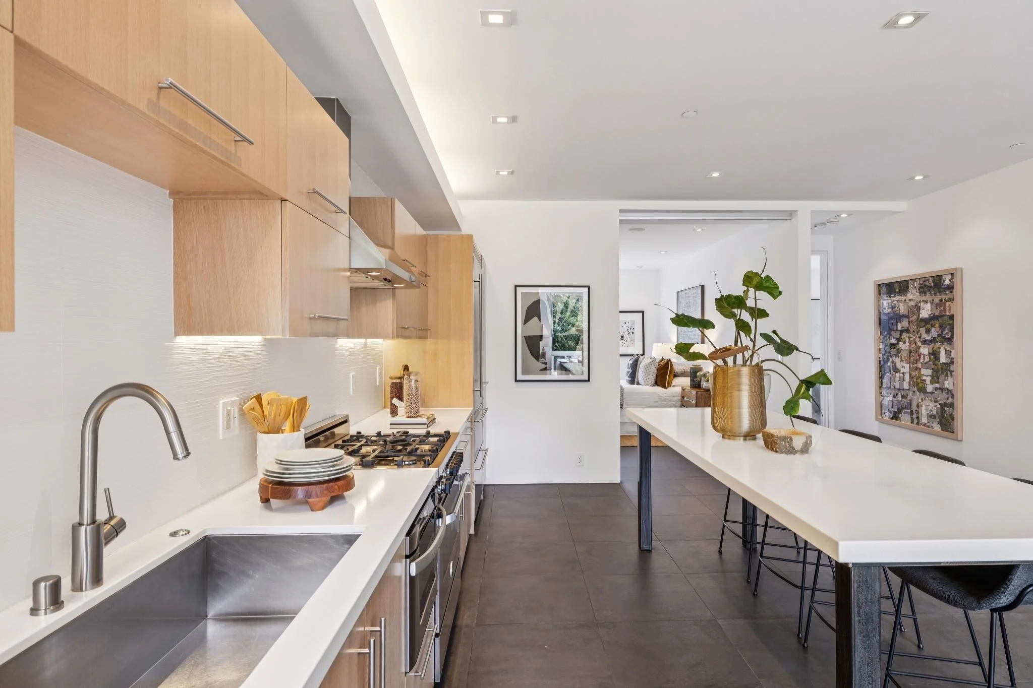 Modern kitchen with wooden cabinets, white countertops, a stainless steel sink, a gas stove, and a dining area with a white table and black chairs, decorated with a large potted plant and artwork on the walls.