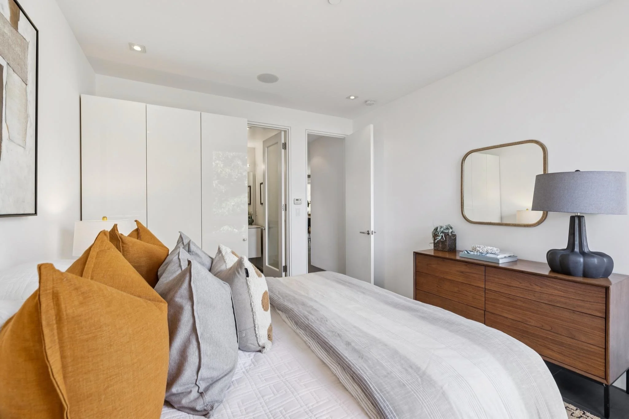 Modern bedroom with white walls, a bed with gray and orange pillows, a wooden dresser with a black lamp, and a rectangular mirror on the wall.