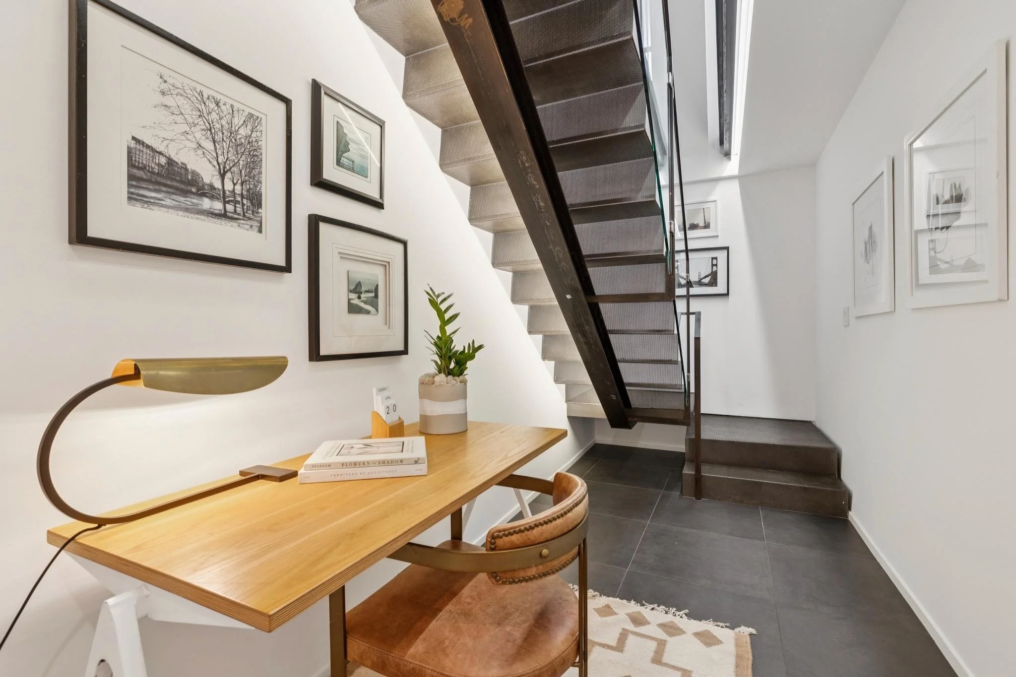 Interior of a modern apartment featuring a wooden desk with a lamp and books, framed pictures on the wall, and a staircase with a dark metal railing leading upstairs.