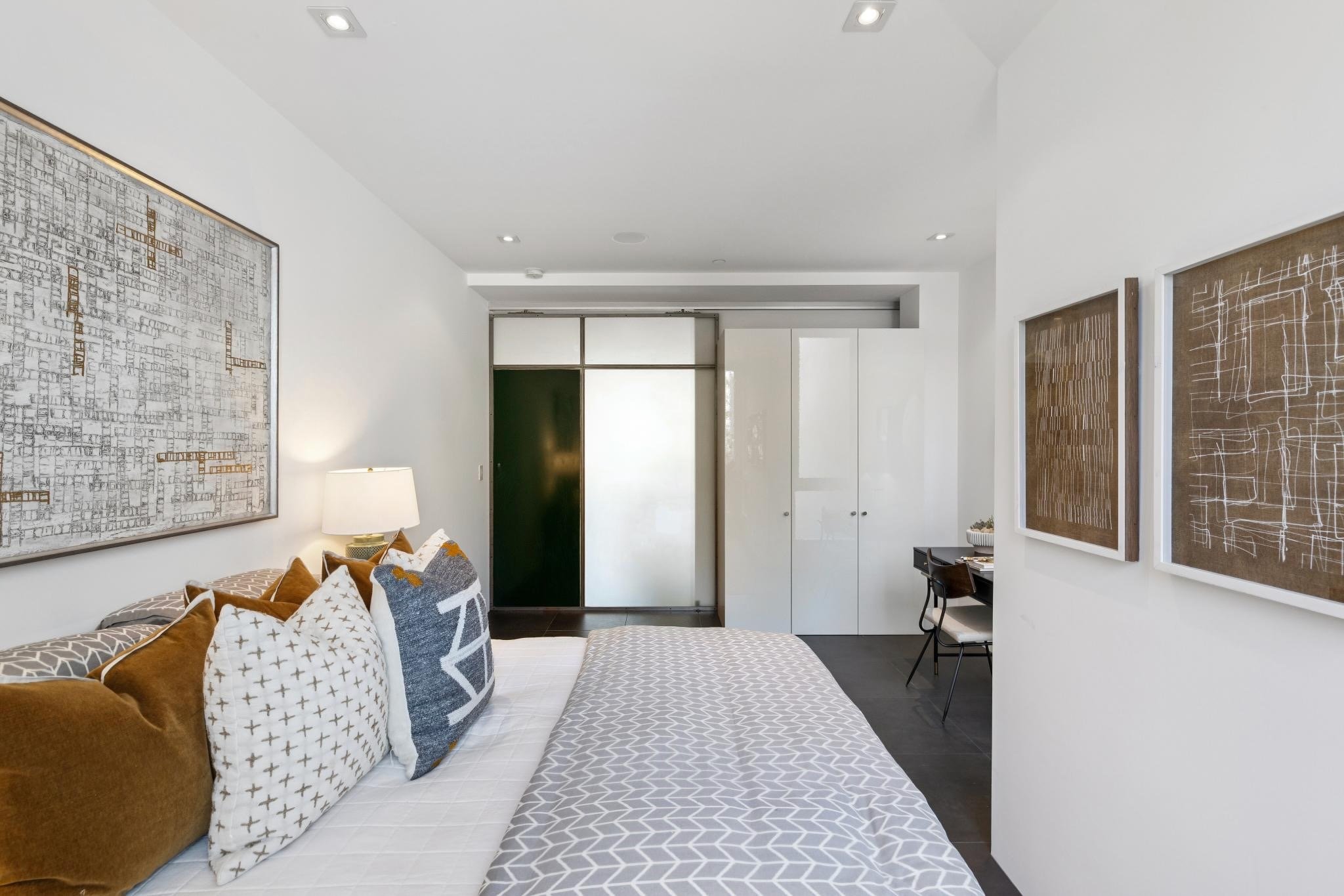 Living room with white walls, framed art, a bed with patterned bedding, pillows, a small desk with a chair, and double doors at the back.