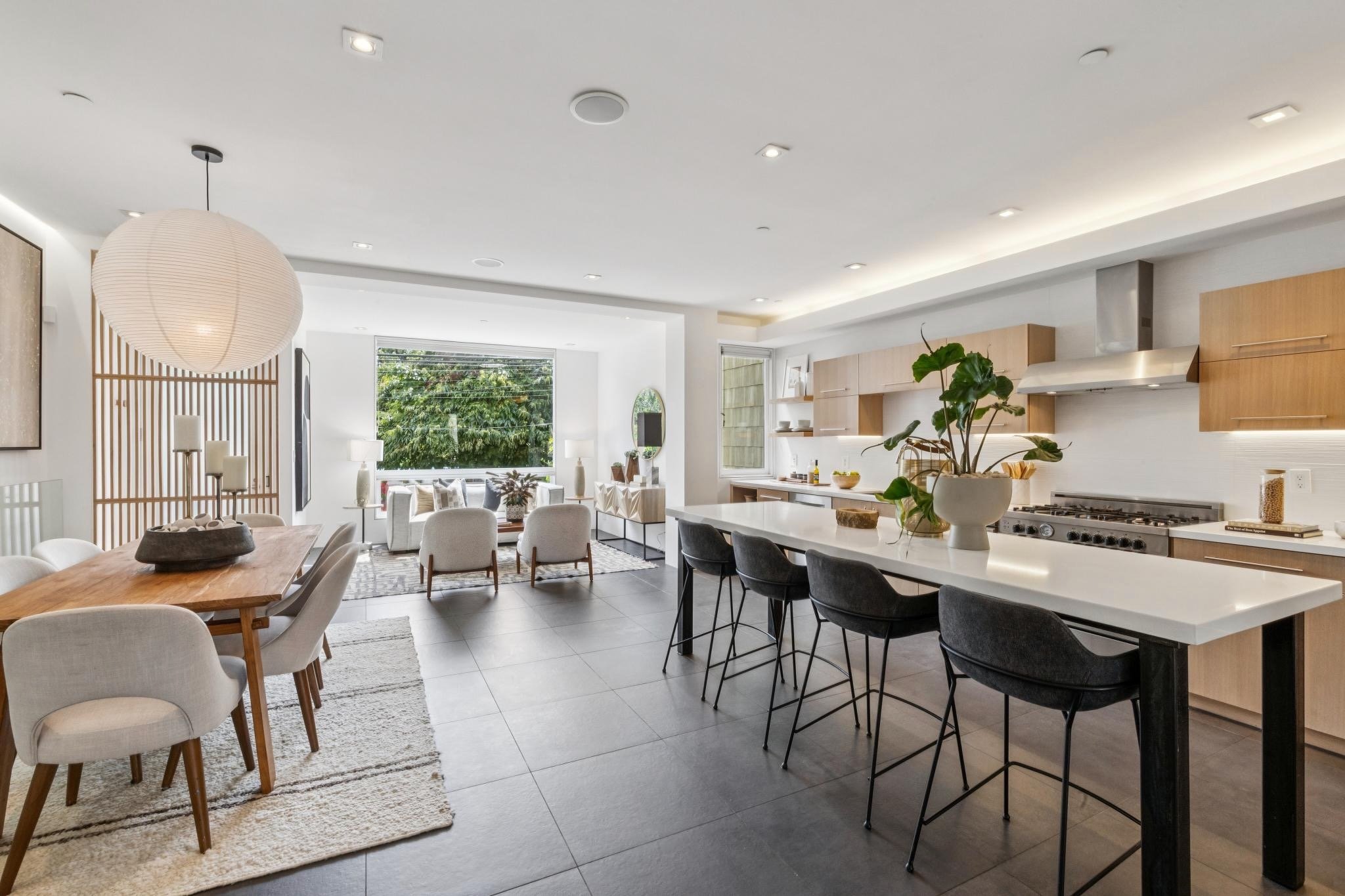 Modern open-concept kitchen and dining area with a large kitchen island, black barstools, light wood cabinets, and a spacious living room with large windows, white walls, and contemporary furniture.