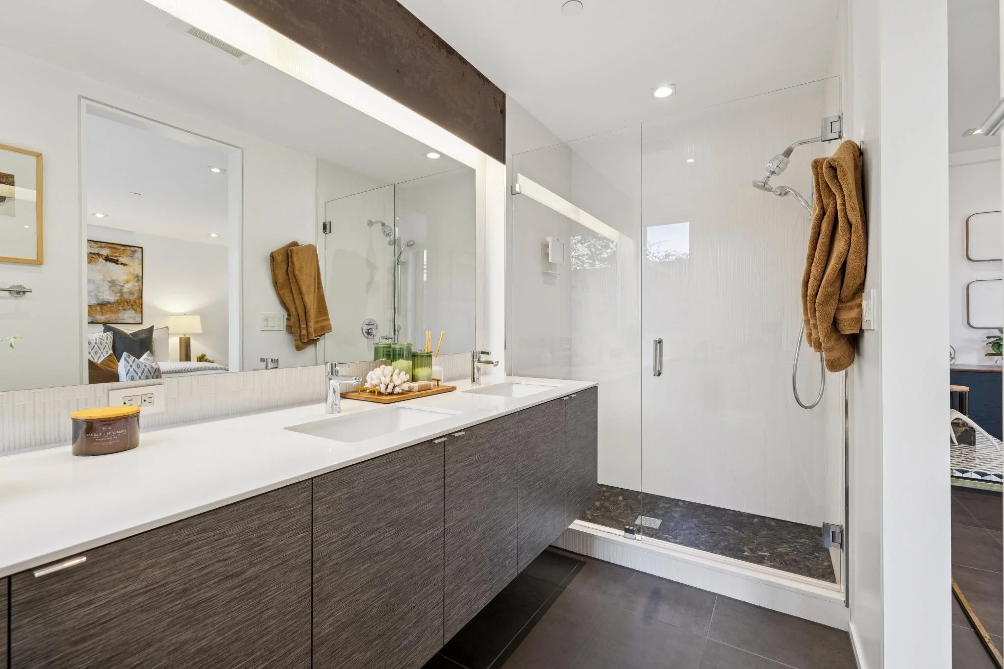View of a modern bathroom with a large mirror, white countertops, and a shower area with glass door and brown towels hanging on the wall.