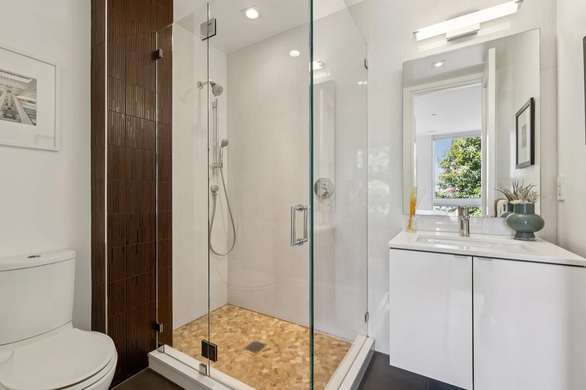 A modern bathroom with a glass-enclosed shower, a white vanity with a sink, a window with outdoor greenery, and a toilet. The shower has a handheld showerhead and wood-textured tiles on one wall. There are framed pictures on the white walls and a dec