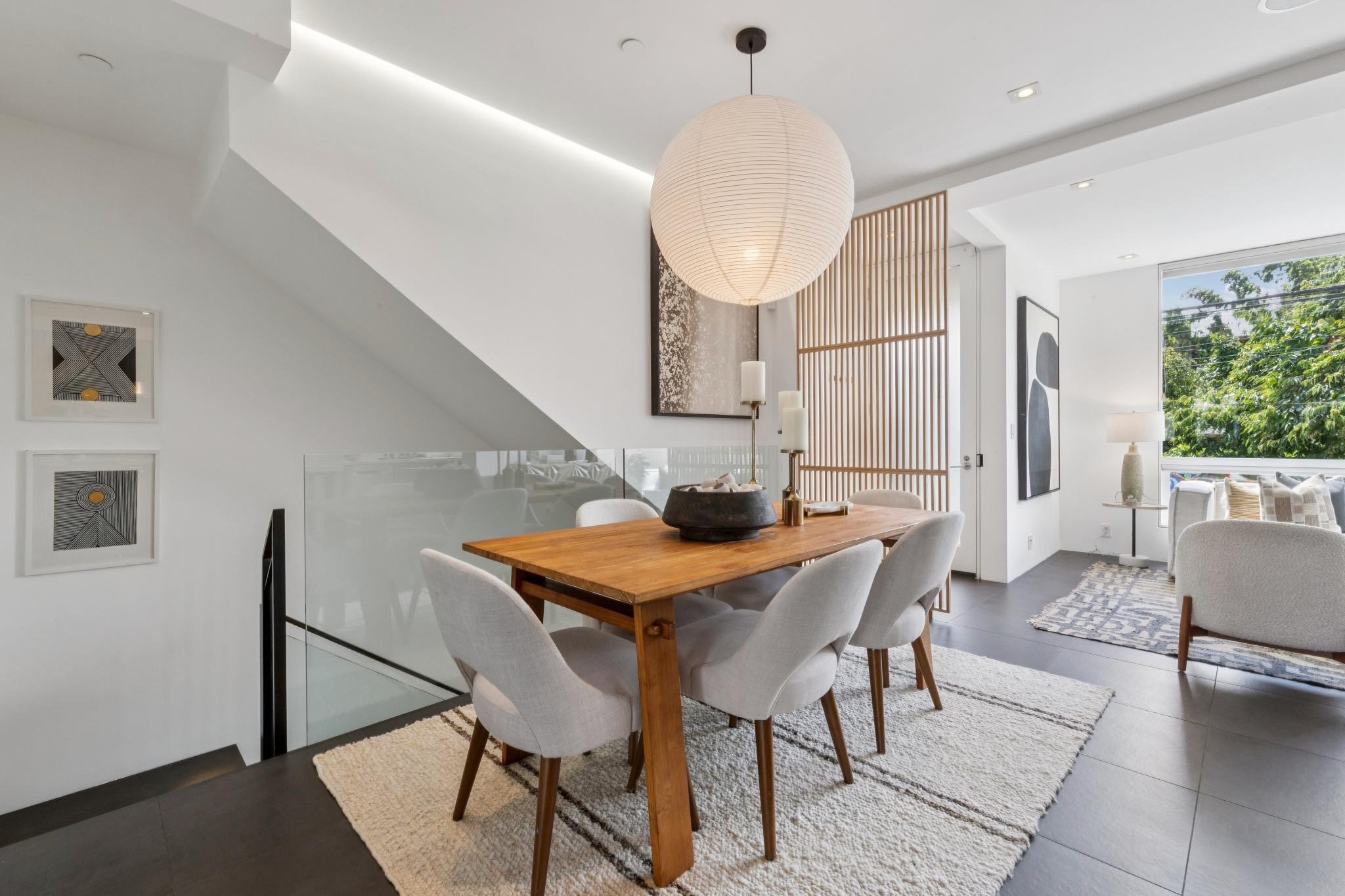 A modern dining room with a wooden table surrounded by eight upholstered chairs on a patterned area rug. The room has white walls with framed abstract art, a large paper lantern pendant light, and large windows showing greenery outside.