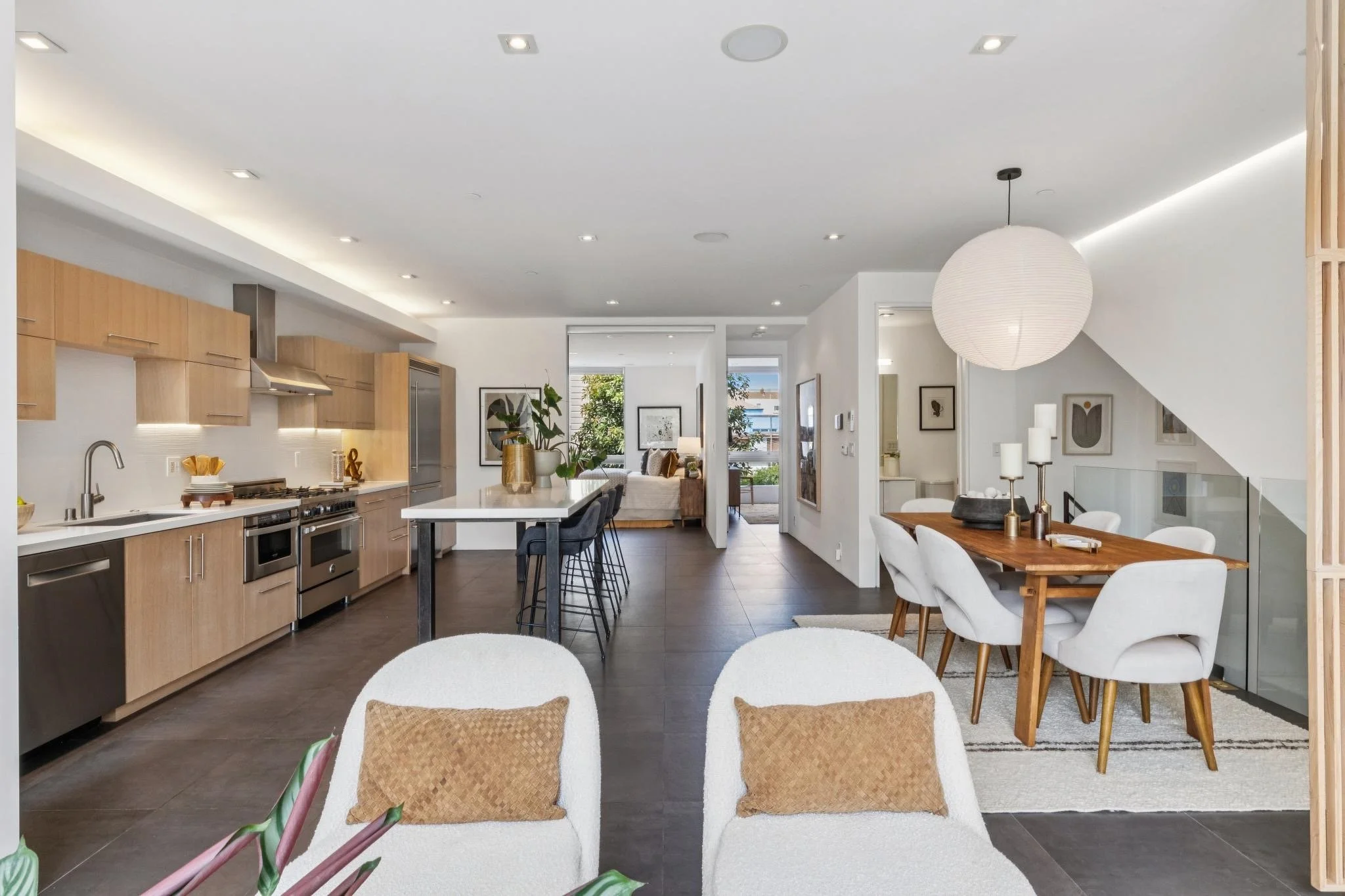 Open-concept kitchen and dining area with white walls, wooden cabinetry, modern appliances, and a large dining table with white chairs, decorated with a pendant light, candles, and artwork.