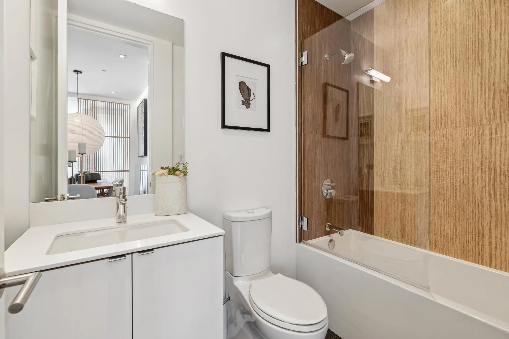 A modern bathroom with a white vanity, a framed abstract art piece on the wall, a white toilet, and a glass shower with wooden panels inside.