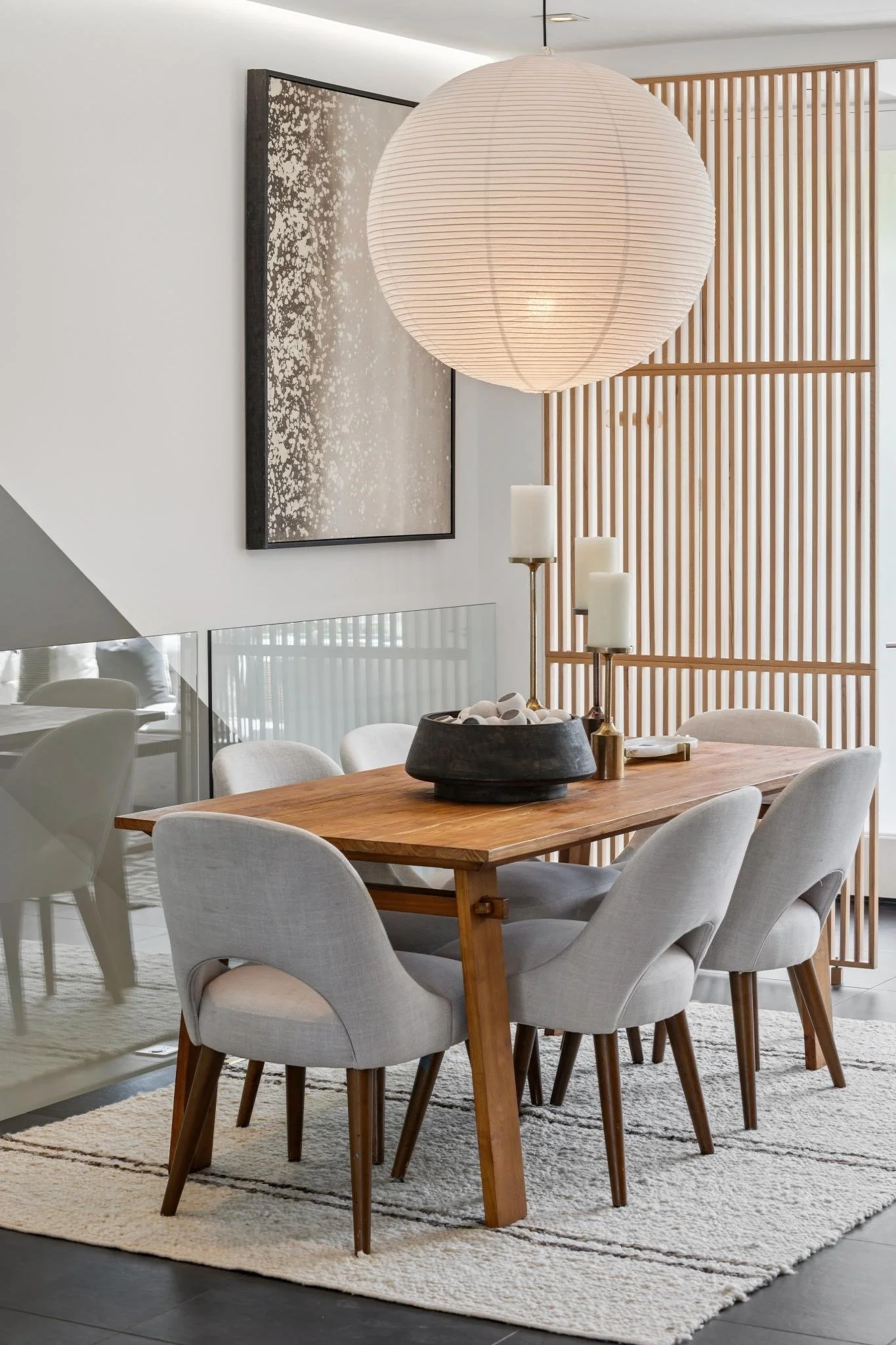 A dining room with a wooden table surrounded by beige upholstered chairs with wooden legs. A large white paper lantern hangs from the ceiling, and a black container with decorative stones and three candle holders with white candles are on the table. 