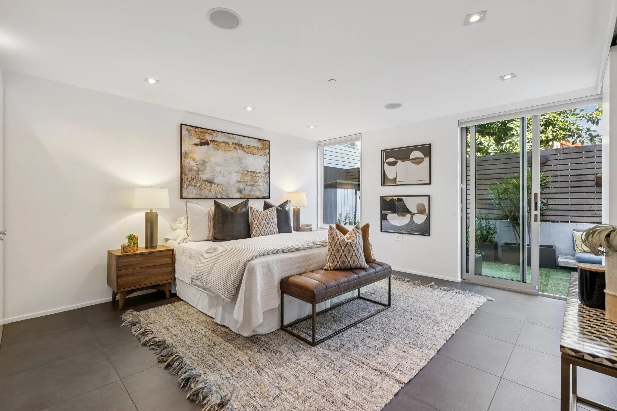 A modern bedroom with a bed, pillows, framed artwork, a sliding glass door opening to a small outdoor patio, and a rug on dark tile flooring.